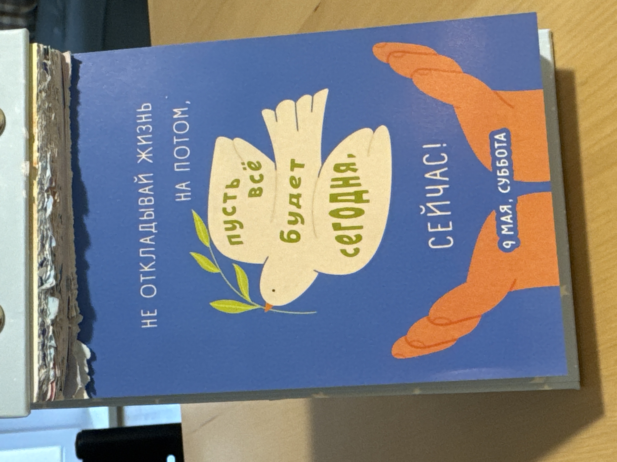 Here's a forensic description of the image:
The image shows a desk calendar, open to a page with a blue background. The text on the page is written in Russian, and the illustration includes a white dove with an olive branch, surrounded by orange hands. The text reads "НЕ ОТКЛАДЫВАЙ ЖИЗНЬ НА ПОТОМ," which translates to "Don't put off life until later." The next lines, "пусть всё будет сегодня," translates to "let everything be today," and "СЕЙЧАС!" means "NOW!" Below the hands it reads, "9 МАЯ, СУББОТА," which translates to "May 9th, Saturday". The location of the image is likely an indoor space. There is no indication of Люк or Russia.
