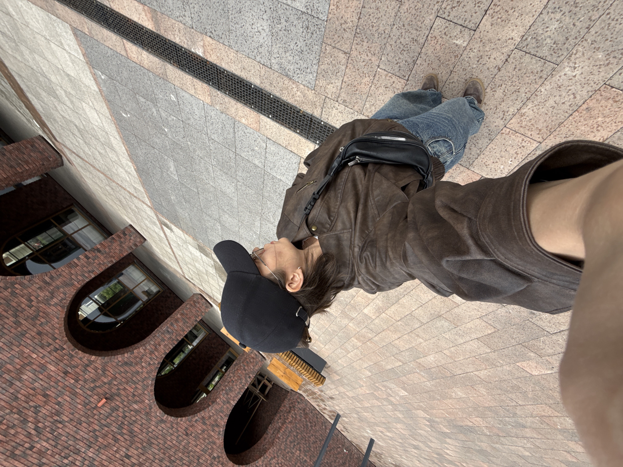A high-angle, slightly rotated (90 degrees clockwise) overhead shot captures a person standing outdoors in what appears to be a paved urban area in Izhevsk, Russia. The individual, seen from behind and slightly to the side, is wearing a brown leather jacket, blue jeans, and a black baseball cap. A black fanny pack or crossbody bag is worn over the jacket. The person also has glasses and a small earring visible on their left ear.
The ground beneath the person is a mosaic of different colored and textured paving stones, predominantly grey and light brown, arranged in geometric patterns. A long, black metal drainage grate runs across the paved surface. To the left side of the frame, a modern brick building with distinctive arched windows is visible. The brickwork is reddish-brown. Reflections of trees or greenery can be seen in some of the window panes.
The lighting suggests it is daytime, with soft, diffused light, indicating either an overcast sky or an indirect light source, as no harsh shadows are cast. There is no visible text in the image, and the person's emotions are not discernible from this perspective. The scene depicts a calm, everyday urban environment.