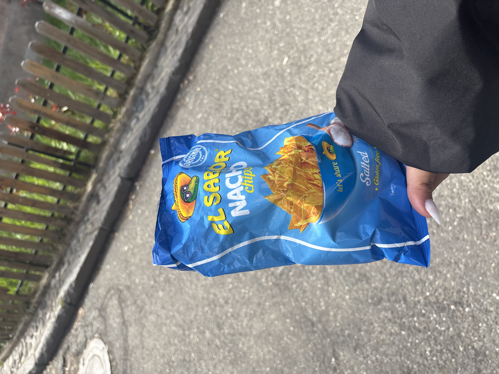 A person in a black jacket holds a bag of "El Sabor Nacho Chips" in their right hand. The bag is blue with a picture of tortilla chips on it. The words "El Sabor Nacho Chips" are prominently displayed in yellow. The bag also indicates that the chips are "Salted" and "Gluten Free." The location appears to be outdoors on a sunny day, with a sidewalk or paved area visible. A fence line borders the edge of the image. The city and country are not discernible from the visual information available.