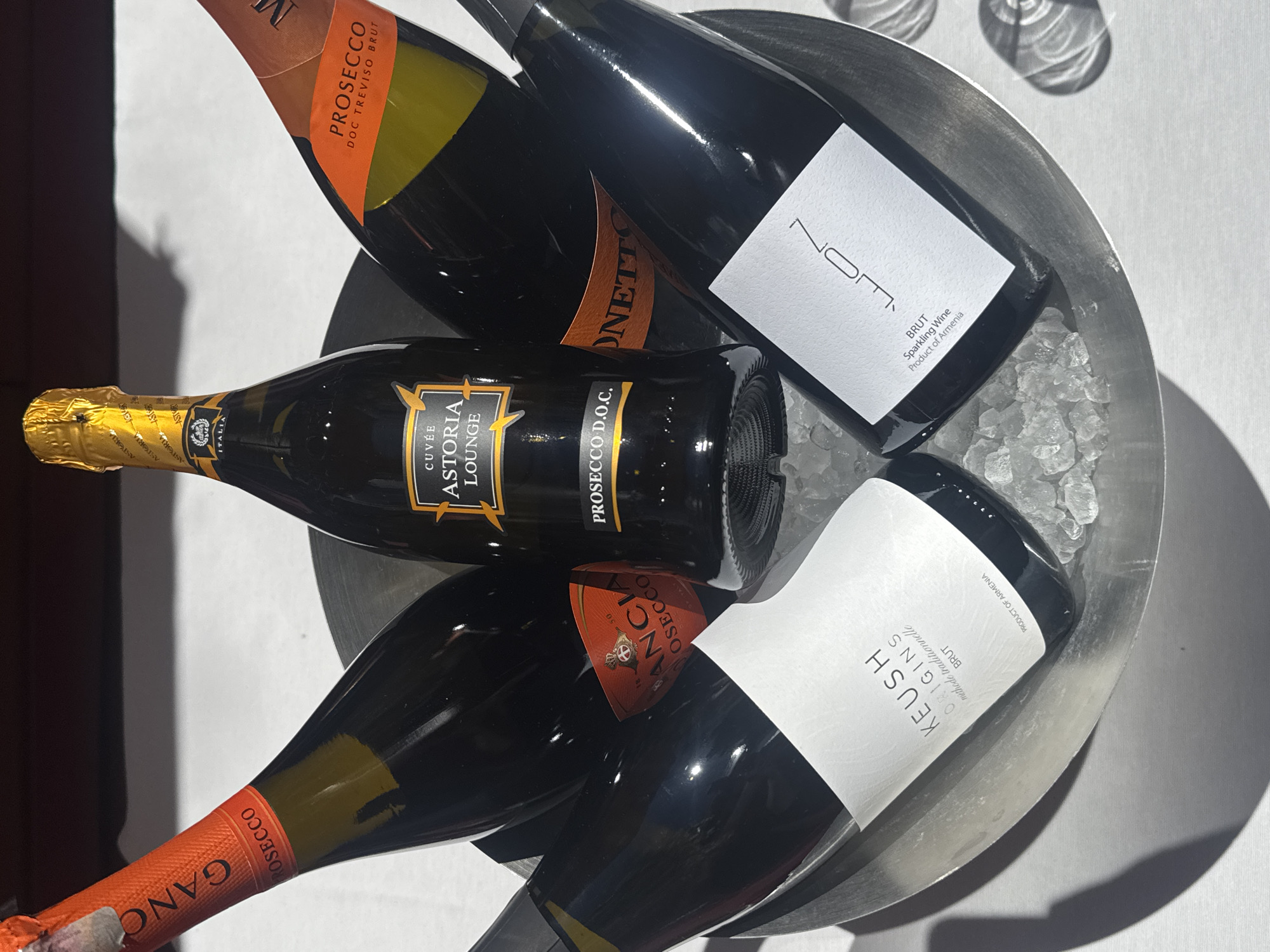 A low-angle, medium shot depicts a silver ice bucket containing five bottles of sparkling wine or prosecco. Labels on the bottles include "GANCIA PROSECCO," "KEUSH ORIGINS BRUT" (Product of Armenia), "CUVÉE ASTORIA LOUNGE PROSECCO D.O.C.," "NOM BRUT Sparkling Wine Product of Armenia," and "DONETTO PROSECCO DOC TREVISO BRUT." The bucket contains ice and sits on a white surface with shadows nearby. No people are visible in the image. The location is likely Yerevan, Armenia, given the mention of Armenia on two of the bottle labels.