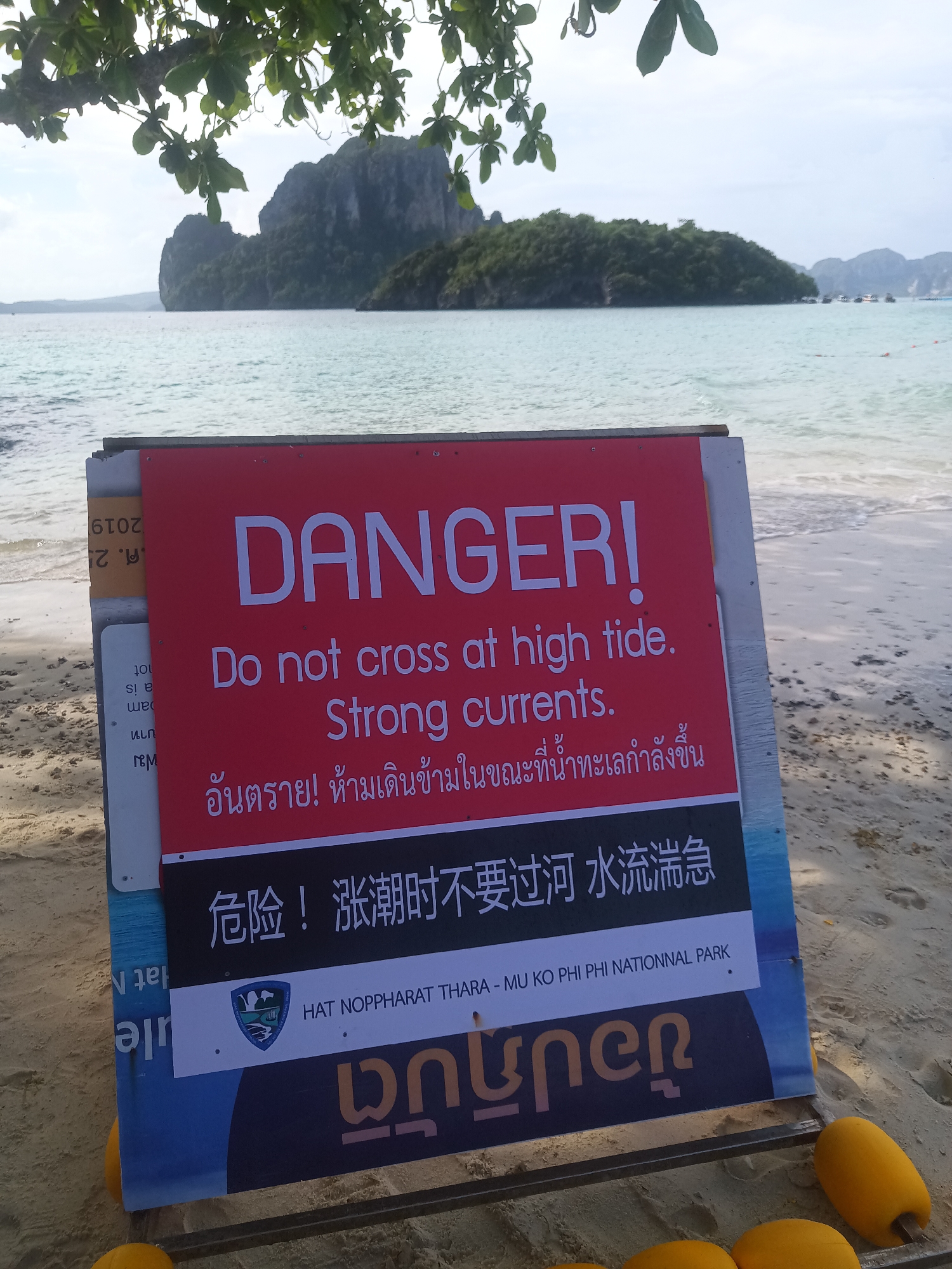 The image is a beach scene featuring a warning sign placed on the sand. The sign is predominantly red and white, with "DANGER!" prominently displayed in large, bold letters. Below this, the sign instructs, "Do not cross at high tide. Strong currents." The warning is translated into Thai and Chinese below the English text. The sign also includes a logo for Hat Noppharat Thara - Mu Ko Phi Phi National Park and a text in an unknown language. The sign is surrounded by yellow safety buoys. Behind the sign, there is a tropical beach with light sand, turquoise water, and an island with rocky formations covered in green vegetation. Some boats are visible in the distance, as is a tree.