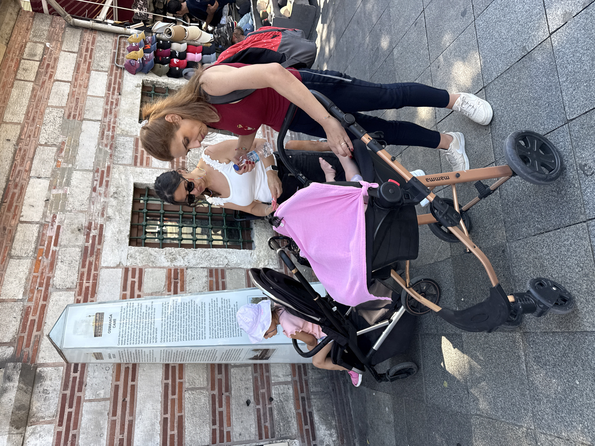 In front of a brick and stone building with barred windows, two women and a young child are near strollers on a paved street. The child, wearing a pink dress and hat, sits in a black stroller. One woman, in a red tank top and dark pants, tends to the stroller, holding a water bottle. The other woman, in a white top and dark pants, stands beside her. A market stall with hats is visible in the background. The building features a tall, white informational display. The scene is likely near the Osmanaga Mosque in Kadıköy, Turkey, given the inscription on the informational display.