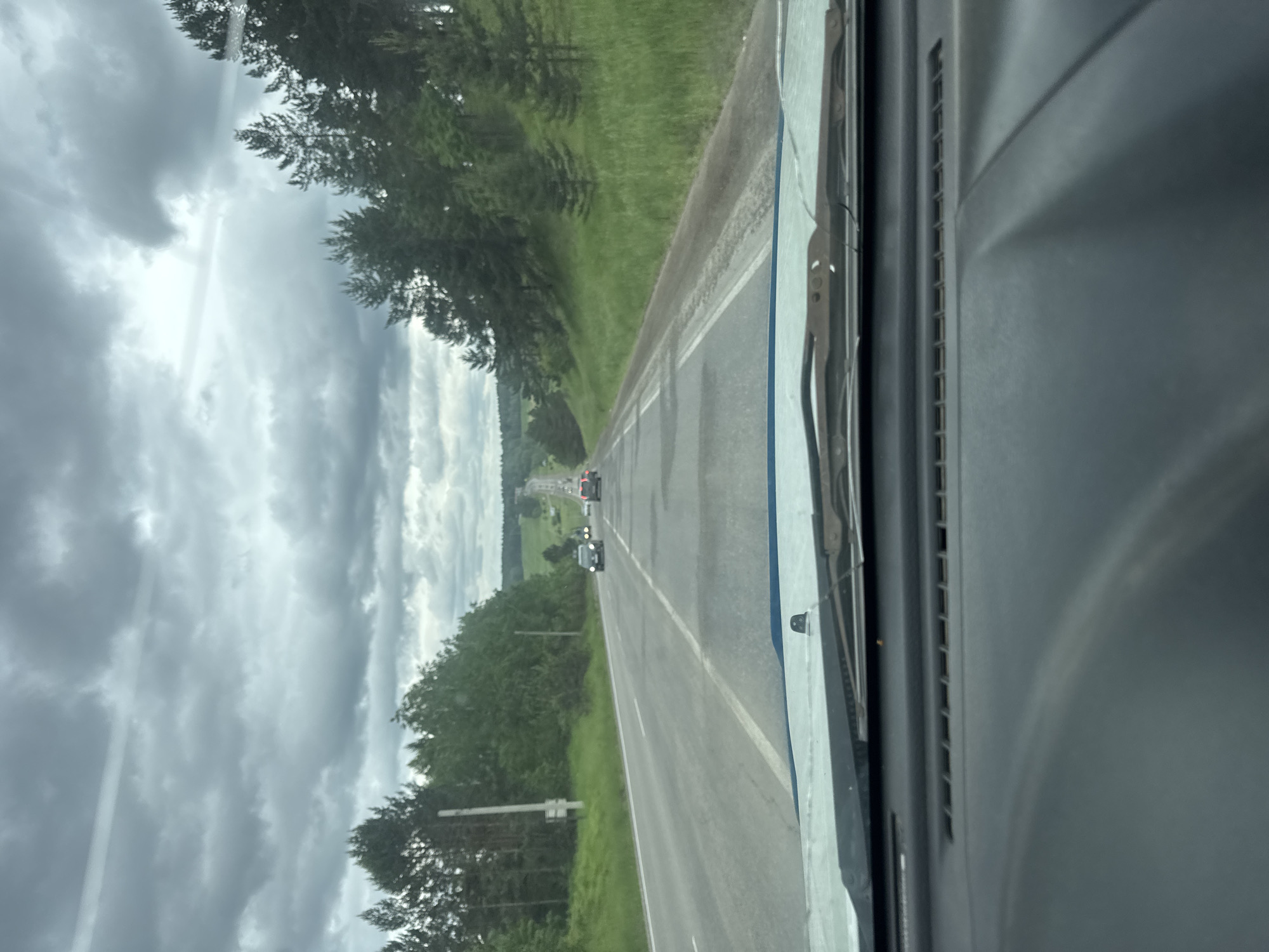 The image shows a view from inside a vehicle driving on a rural road near Gornyy, Russia. The road is paved and has multiple lanes, with several cars in front. The landscape is characterized by rolling green hills, dense forests, and a cloudy sky. The weather appears overcast, with dark clouds dominating the sky. The road surface is uneven, with visible patches and signs of wear. The vegetation is lush and green, indicating a temperate climate. The scene depicts a typical journey through a rural area, with a focus on the natural environment and the road infrastructure.