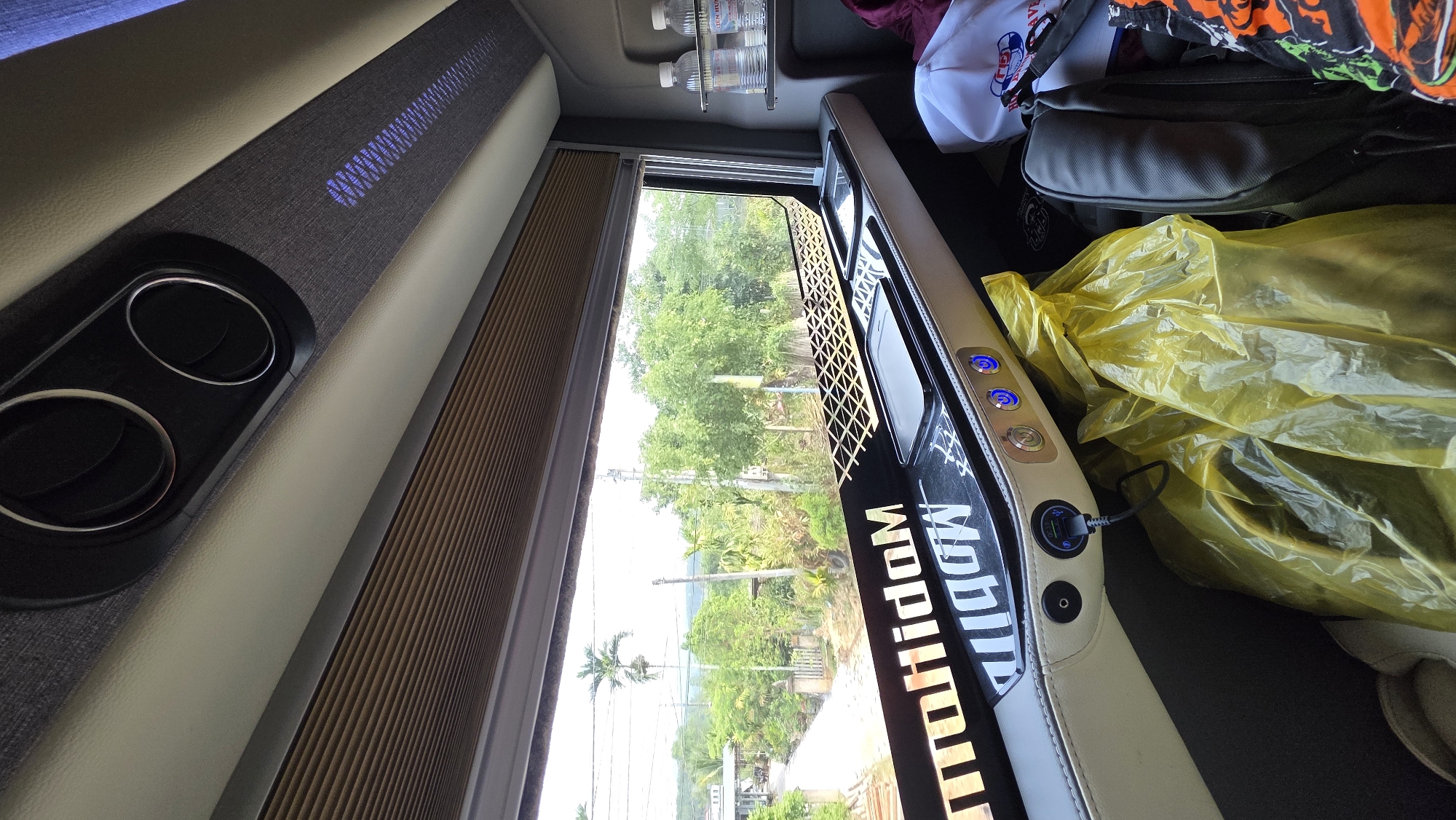 The scene is an interior view from a bus or specialized vehicle, likely in Dak To, Vietnam. The interior features include customized trim with illuminated lettering, possibly indicating the brand "Mobile", along with controls for lights or other functions. A window shows an exterior landscape with trees, a road, and utility poles. Luggage or bags and plastic yellow bag containing a personal item are piled on the seat. Inside the bus are bottle of water in a metal holder.