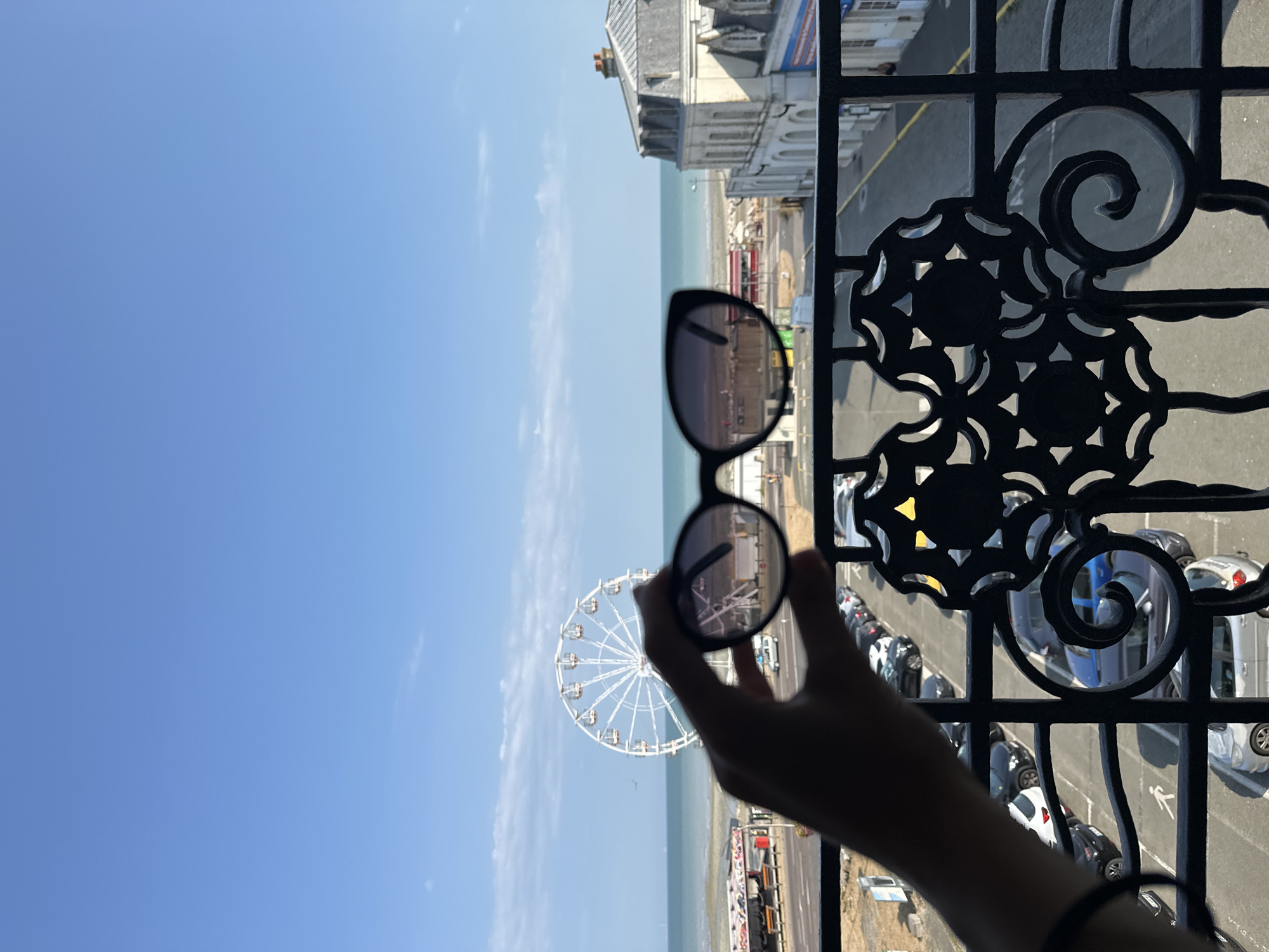 The image shows a view from a balcony overlooking a street and a coastline in Le Tréport, France. In the foreground, a hand holds up a pair of sunglasses, obscuring part of the view. Through the sunglasses, a Ferris wheel and the sea are visible. The background features a street with parked cars and buildings along the coast, typical of a seaside resort town. The sky is clear and blue, suggesting a sunny day.