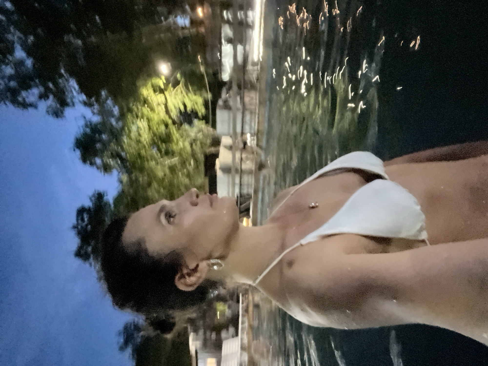 A woman is partially submerged in a body of water at dusk. She is wearing a white bikini and looking to her right. The water is dark with reflections of lights and trees. In the background, there are trees and some structures that appear to be part of a resort or spa, possibly indicative of the Paratunka region of Russia, known for its hot springs and recreational facilities. The sky is a deep blue, suggesting it is either twilight or night.