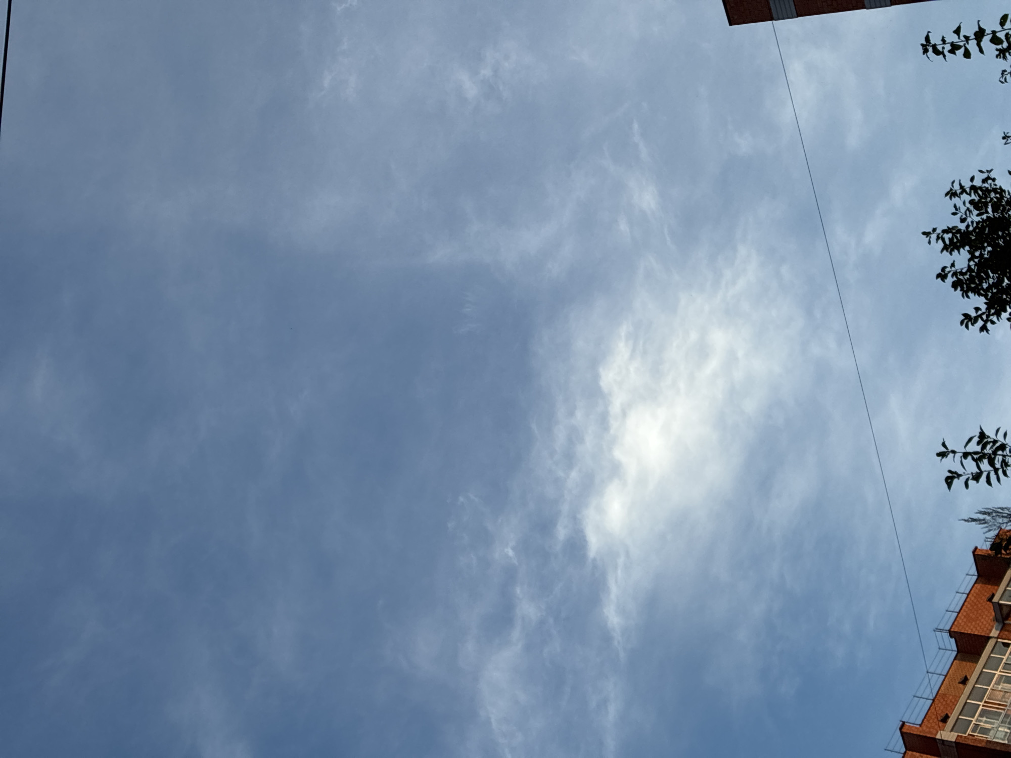 The image shows a bright blue sky with scattered wispy white clouds. In the lower left corner, the top of a brick building with windows is visible, and a black wire stretches horizontally across the sky. Silhouettes of tree branches are visible in the lower portion of the image, against the sky. The scene appears to be an outdoor urban environment, possibly in Markova, Russia, given the context.