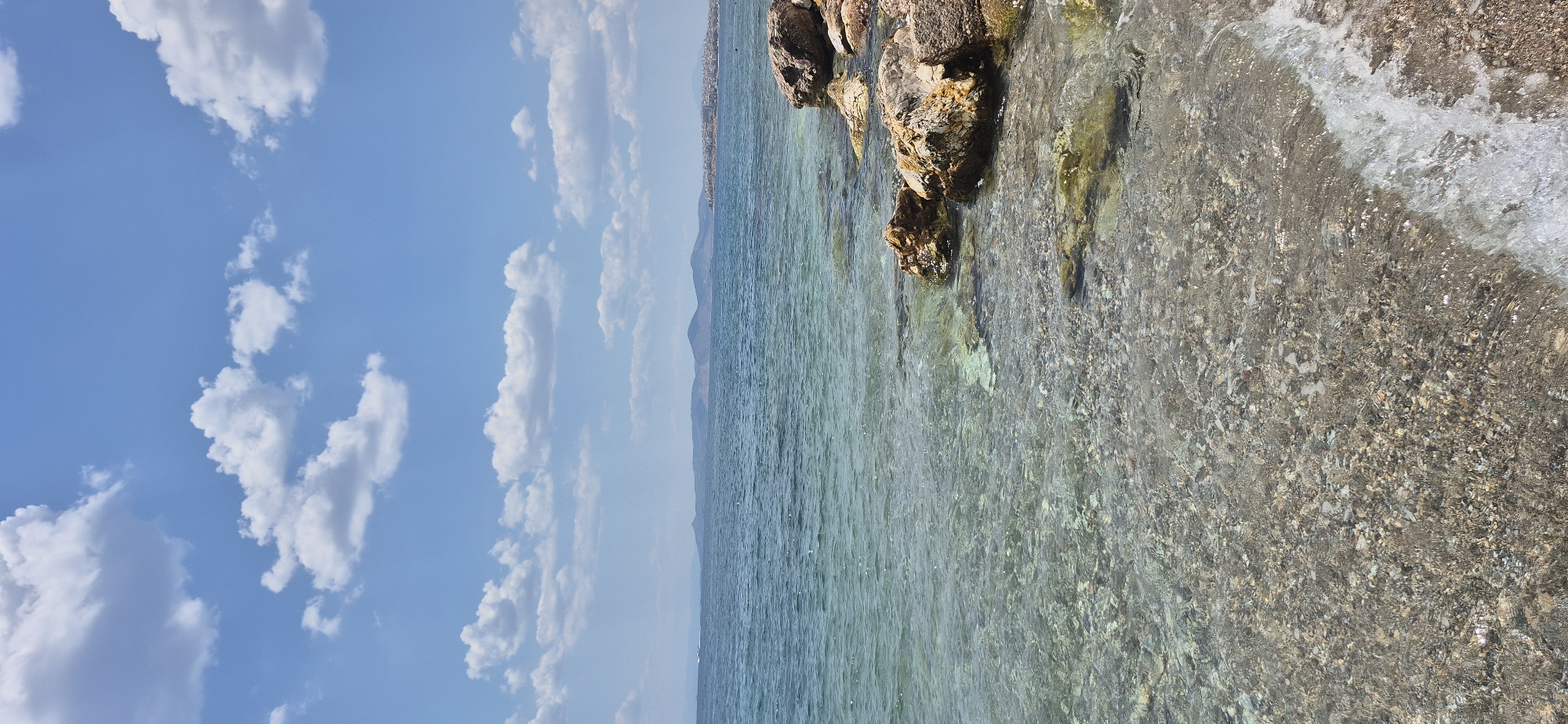 This media file depicts a scene of a clear, calm sea bordering a rocky shore, under a bright blue sky dotted with fluffy white clouds. The image is oriented vertically.
The subjects include the natural elements of the environment: the sea, rocks, and sky. The sea water is remarkably transparent, revealing the pebble-strewn seabed near the shore. Larger rocks, varying in color from brown to reddish-brown, are present at the water's edge, some partially submerged. The sky is a vibrant blue, with several cumulus clouds scattered across it. In the distance, a hazy landmass with a mountainous silhouette is visible, suggesting a coastline or island. A faint line of structures, possibly buildings, can be seen along this distant shore.
No people or specific objects are distinctly visible. The scene suggests a peaceful and serene setting, likely a beach or coastal area. The lighting indicates it is daytime, with the sun casting a slight shimmer on the water's surface. The weather appears to be pleasant and sunny.
The location context of Paleo Faliro, Greece, suggests this is a Mediterranean coastal scene. The clear water and rocky shore are consistent with many Greek coastlines. No text is visible in the media file. The overall emotion evoked is one of tranquility and natural beauty.