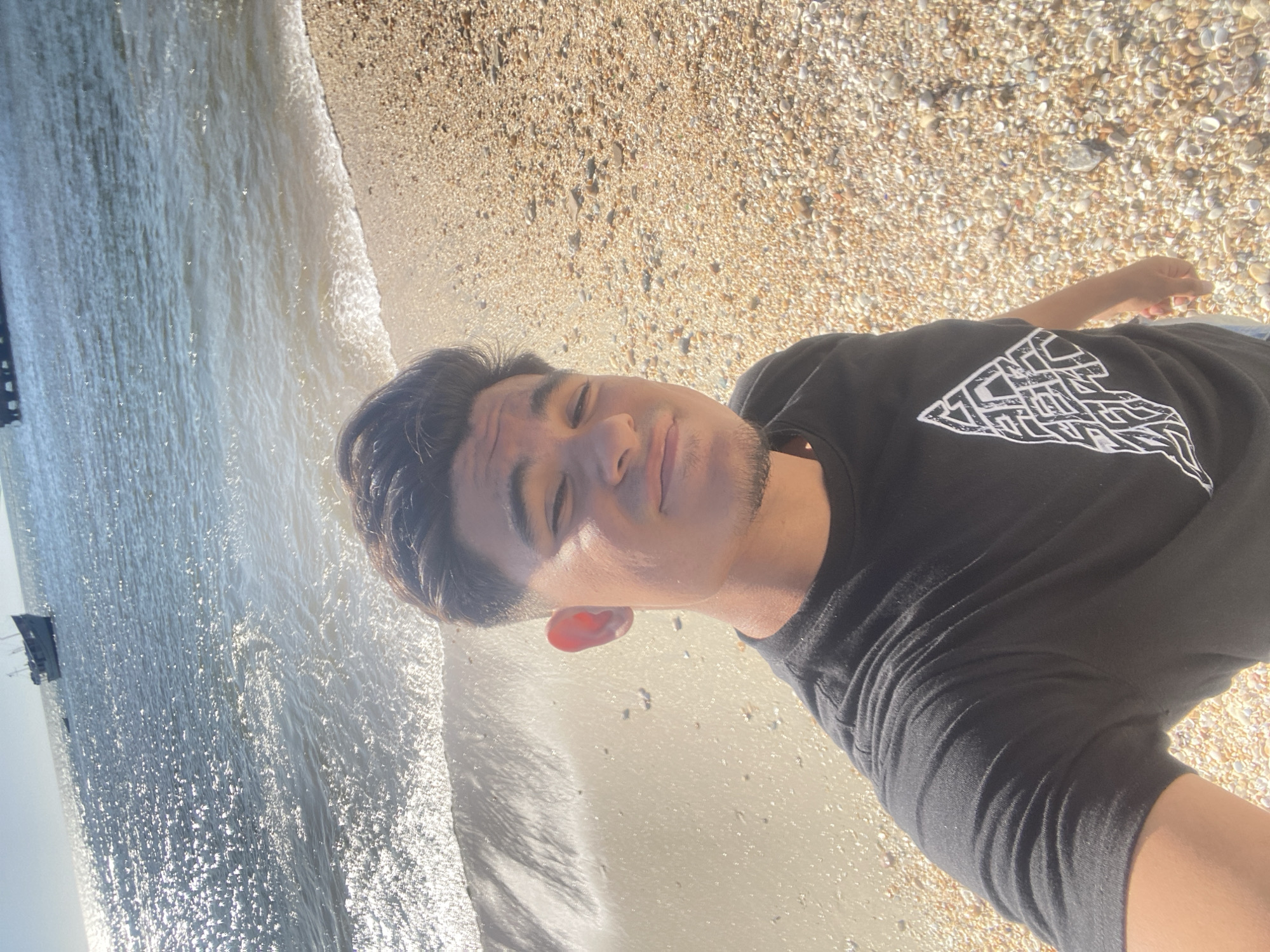 The image captures a male individual taking a selfie on a beach in Juan Griego, Venezuela. The man, positioned in the foreground, has dark hair, light brown skin, and a visible goatee. He is wearing a black short-sleeved t-shirt with a distinct white, triangular graphic design on the left chest. He is smiling faintly and looking directly at the camera. The immediate environment consists of a light-colored sandy beach transitioning to an area of small, mixed-color pebbles and seashells further from the water's edge. The ocean occupies the background, displaying sun glare across its surface, particularly on the left. Small waves are breaking at the shoreline. Two watercraft are visible in the distant ocean: a light-colored, multi-decked vessel with a mast structure on the left, and a less defined dark structure or vessel on the right, possibly a pier. The scene is illuminated by bright daylight.