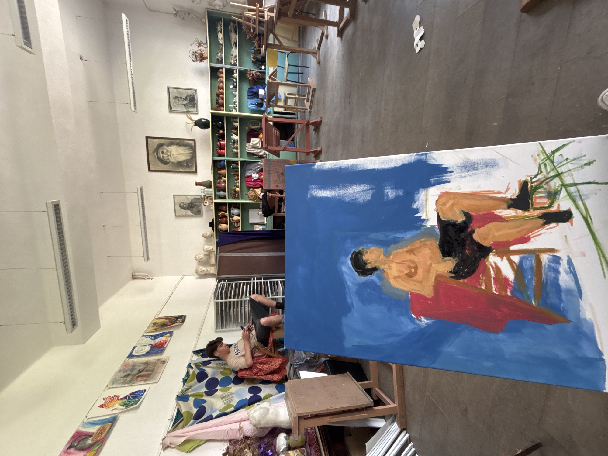 An art studio in Troyan, Bulgaria, features a model reclining on a colorful patterned cloth on the floor, using a phone. A painting on an easel depicts a similar figure against a blue background. The studio is filled with art supplies, paintings, and shelves filled with objects.