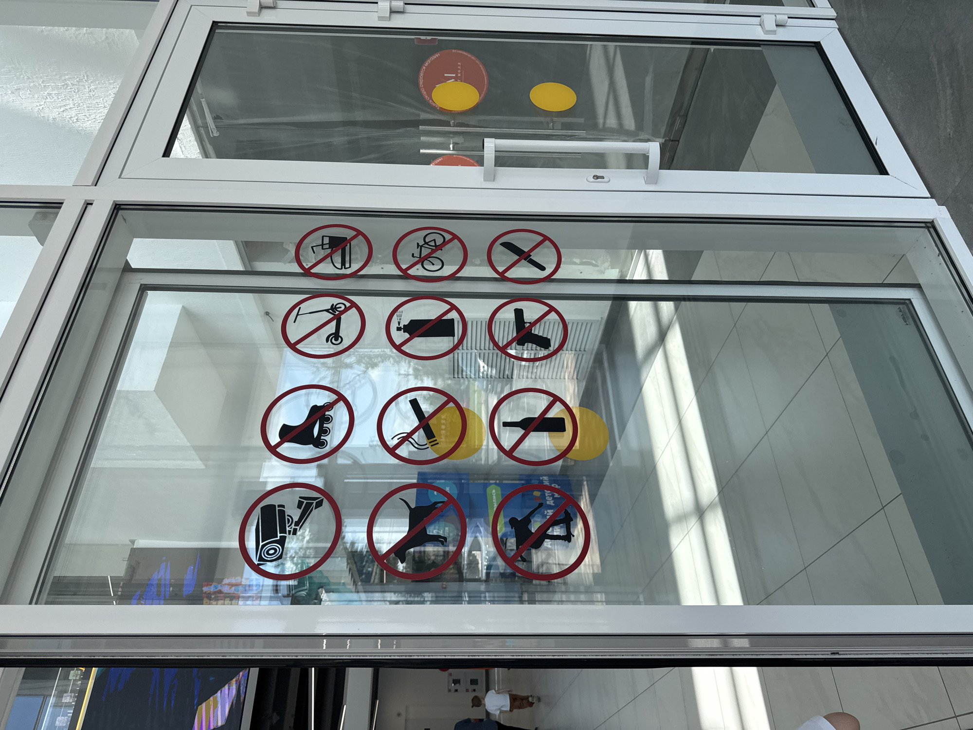 The image shows a glass entrance with several "no" signs. These signs, displayed in red circles with a diagonal line, prohibit a variety of activities and items. They include a security camera, roller skates, a scooter, a burger and drink, a dog, a cigarette, a bicycle, spray paint, a handgun, a knife, and a bottle. The glass reflects the interior and exterior of the building.
The location is the entrance to the "Galereya" shopping mall in Mikhaylovsk, Russia. The mall's logo is visible on the interior glass door. Two yellow circular stickers, likely indicating floor-level obstacles or safety markings, are also present on the interior glass.
In the background, a young boy in a white t-shirt and brown pants is walking away from the camera down a tiled hallway. The hallway is brightly lit, with sunlight streaming through windows or skylights, creating geometric patterns of light and shadow on the floor. The overall environment appears to be a modern, clean, and well-maintained public space.