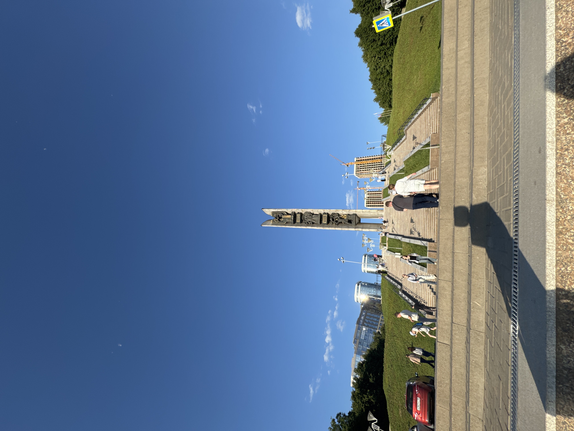 The image displays a wide-angle view of the Friendship of Peoples Monument and its surrounding area in Izhevsk, Russia, under a clear blue sky.
The central feature is a tall, vertical monument, constructed of light-colored material with dark, intricate bas-reliefs on its sides, situated at the top of a large, multi-tiered concrete staircase. This monument stands centrally in the upper midground.
In the foreground, wide, shallow concrete steps are visible, followed by a paved section featuring a metal drainage grate. A prominent, elongated dark shadow is cast diagonally across these foreground steps from the lower left towards the right.
The midground is dominated by the extensive concrete staircase, which ascends a green grassy slope. Multiple individuals are scattered across the stairs and surrounding areas. On the lower left, a red sedan (Mazda 3) is parked or moving slowly near the base of the stairs. Several people, including a child in a light-colored outfit, are walking on the lower stairs and adjacent pathways. In the central lower midground, a man in a dark shirt is pushing a dark stroller or cart, followed by a woman in a white two-piece outfit, both facing away from the viewer and beginning to ascend the stairs. On the right side of the stairs, a blue pedestrian crossing sign with a white walking figure is clearly visible.
The background reveals several modern multi-story buildings with reflective glass facades, some catching the sunlight. Two construction cranes, one yellow and one red, are visible behind buildings on the right, indicating ongoing development. A dense line of green trees borders the upper right grassy slope.
The sky occupies the upper two-thirds of the image, appearing a deep, cloudless blue with only a few very faint, wispy white clouds in the upper right. Several small, bright specks are present in the upper sky, particularly on the left. The scene is brightly lit by the sun, casting distinct shadows.