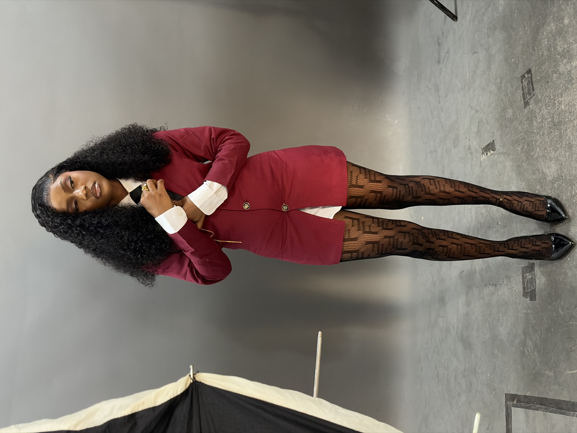 The image displays a full-length, vertically oriented studio portrait of a single young adult female in Gura Top, Nigeria. The woman, with a dark complexion and long, curly black hair, stands facing forward with her head turned to her left, exhibiting a pensive expression with slightly parted, glossy lips. She is dressed in a white collared shirt, a black tie, and a dark red (maroon) blazer-style dress with white cuffs and two visible gold-toned buttons, one fastened. Her hands are clasped in front of her, with her right hand touching her tie, and a gold ring is present on her right index finger. A thin gold chain is visible on her left wrist. She wears sheer black patterned tights featuring a prominent repeating "F" logo (Fendi FF motif) and black patent leather pointed-toe high heels. The background is a plain grey wall and a concrete floor with visible scuff marks and two rectangular dark tape patches. A large black and white photography umbrella on a stand is partially visible on the left side of the frame, with a metallic rod extending from it. A dark stand is also partially visible on the far right, near the floor.