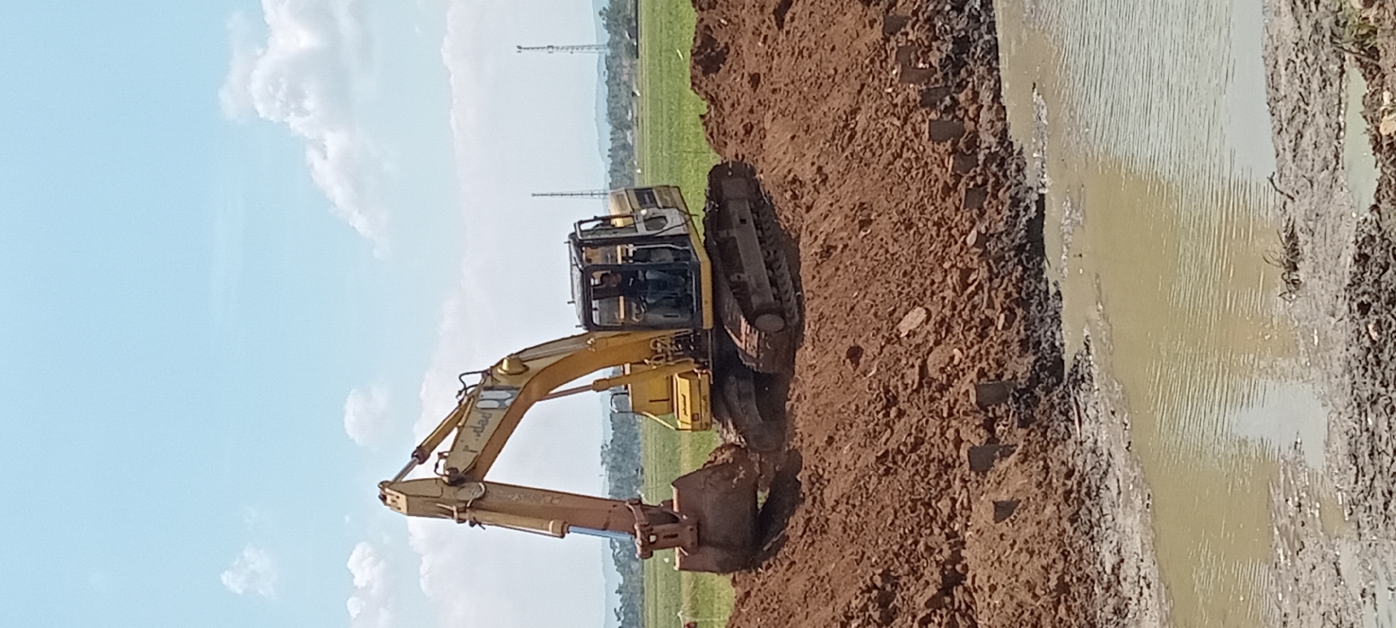 A yellow excavator is pictured in Paledah, Indonesia, on a construction site under a bright, partly cloudy sky. The excavator is positioned on a mound of reddish-brown soil, with its bucket scoop partially submerged in the earth, suggesting it is actively excavating. A single person, wearing a dark shirt, is visible inside the excavator's cab, operating the machinery. In the foreground, a body of murky, light-brown water with ripples covers the ground, indicating a wet or recently flooded area. The background shows a flat, green field with scattered trees and two tall, slender towers, likely communication or utility poles, on the horizon. The scene depicts a construction or land-clearing activity in a rural or semi-rural Indonesian setting.