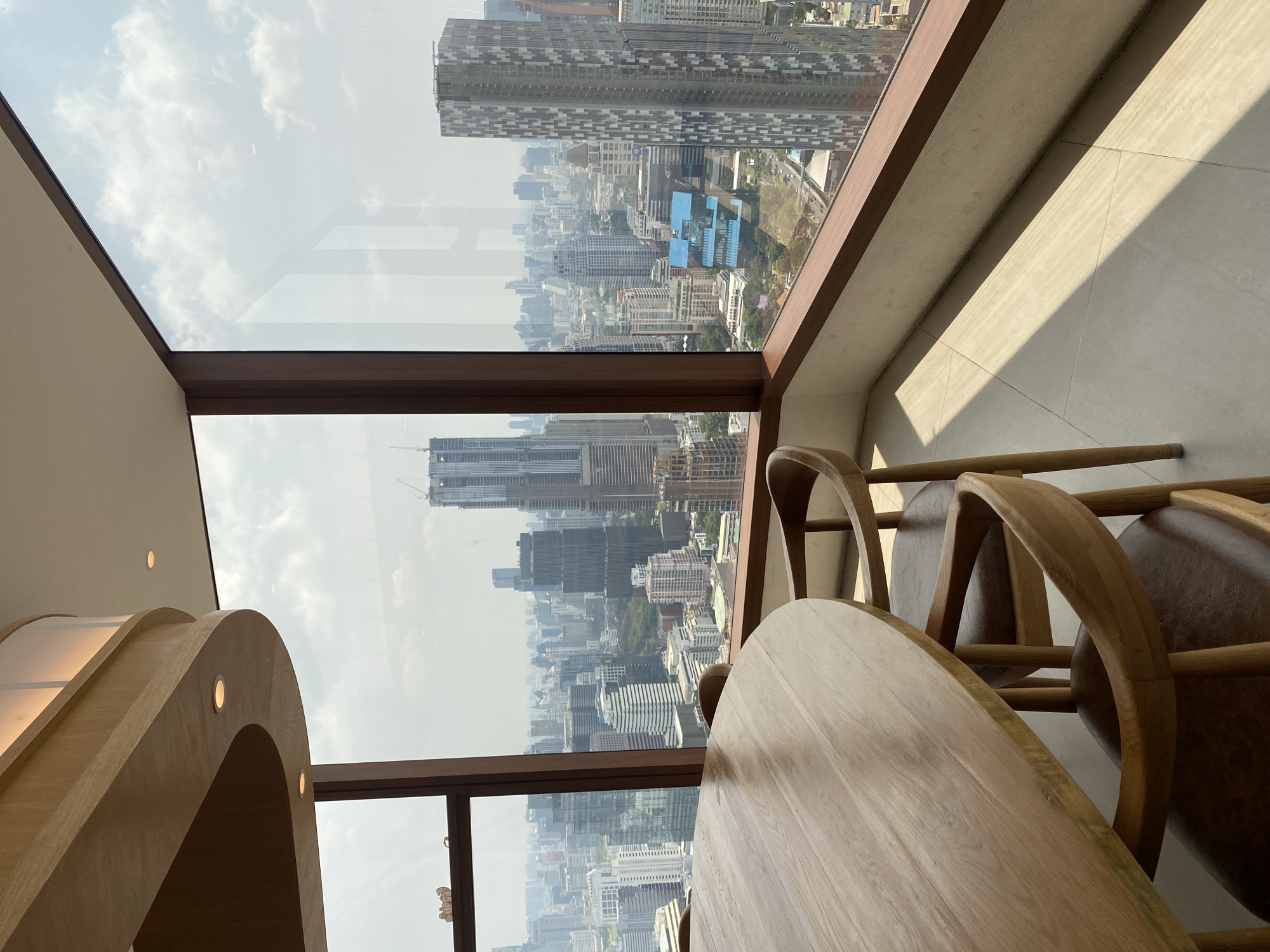 Indoor setting with a view of Bangkok, Thailand. Tall skyscrapers dominate the cityscape visible through a large, multi-pane window. A light-colored wooden table and chairs are in the foreground. Light from the window casts a shadow on the floor. A curved wooden structure with recessed lighting is visible on the left side of the image.