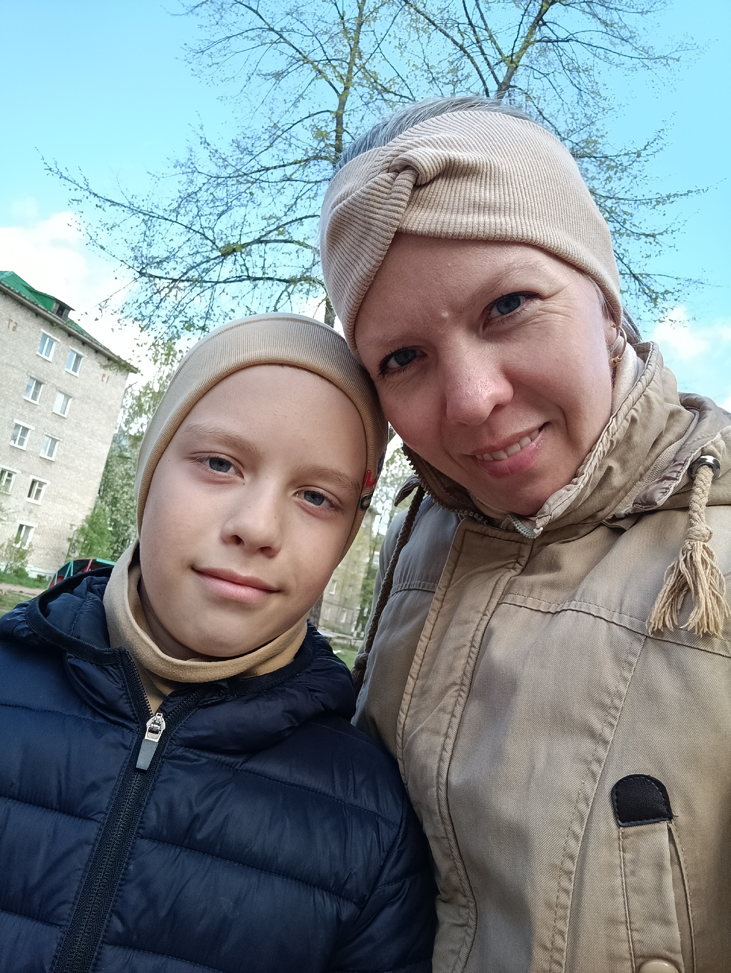 A medium, eye-level outdoor shot depicts a fair-skinned woman and a young boy, likely a mother and son, posing closely for a selfie. The woman, on the right, has a neutral-toned headband, fair skin, and is wearing a tan jacket with tassels hanging from the hood. The boy, on the left, wears a matching neutral-toned beanie and a dark blue, quilted puffer jacket with a high, tan turtleneck. They both have light-colored eyes and are looking directly at the camera with slight smiles.
The background features a light blue sky and budding tree branches. To the left, a multi-story building with rows of windows and a green roof is visible. The photograph's location appears to be in the city of Chaikovsky, Russia, as identified by the metadata. The scene suggests early spring or late autumn due to the clothing and budding trees. The overall composition suggests a casual, personal moment captured in an urban environment.