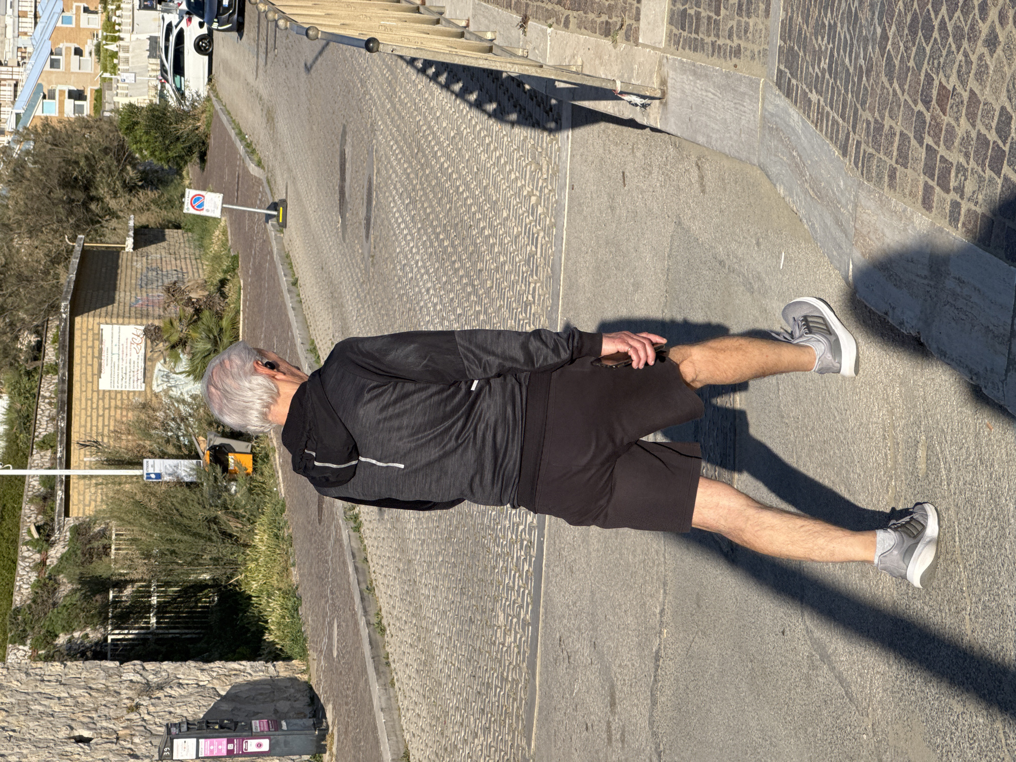 The image, set in Nettuno, Italy, captures a bright, sunny outdoor scene, likely in the morning or late afternoon due to the prominent, elongated shadows cast across the ground. In the center-right foreground, a man with short grey hair is seen from behind, wearing a black long-sleeved athletic top with light stripes on the sleeves, black shorts, and grey and white athletic shoes with white socks. He is engaged in physical activity, stretching by holding his left knee with both hands.
The man stands on a paved surface that combines a patterned, grey cobblestone-like area on the left with a darker, smoother asphalt surface to his right. The ground appears to slope gently, creating a dynamic perspective. Along the right side of the frame, there's a low stone wall topped with a light-colored metal railing.
In the mid-ground on the left, a low-rise building with brick-like walls is partially obscured by lush green shrubs and trees. Some graffiti is visible on its lower section, and a white information board or sign is affixed to it, though its text is unreadable. Further up the road on the left, several other buildings with light-colored facades and pitched roofs are nestled into a hillside, and a white car is parked in front of them.
A red and white "no parking" sign on a pole is visible on the left side of the pathway. In the lower-left corner, a dark grey utility box is partially seen, with the number "39" printed on it. The scene conveys the ambiance of a European coastal town, with clear skies and strong natural light.