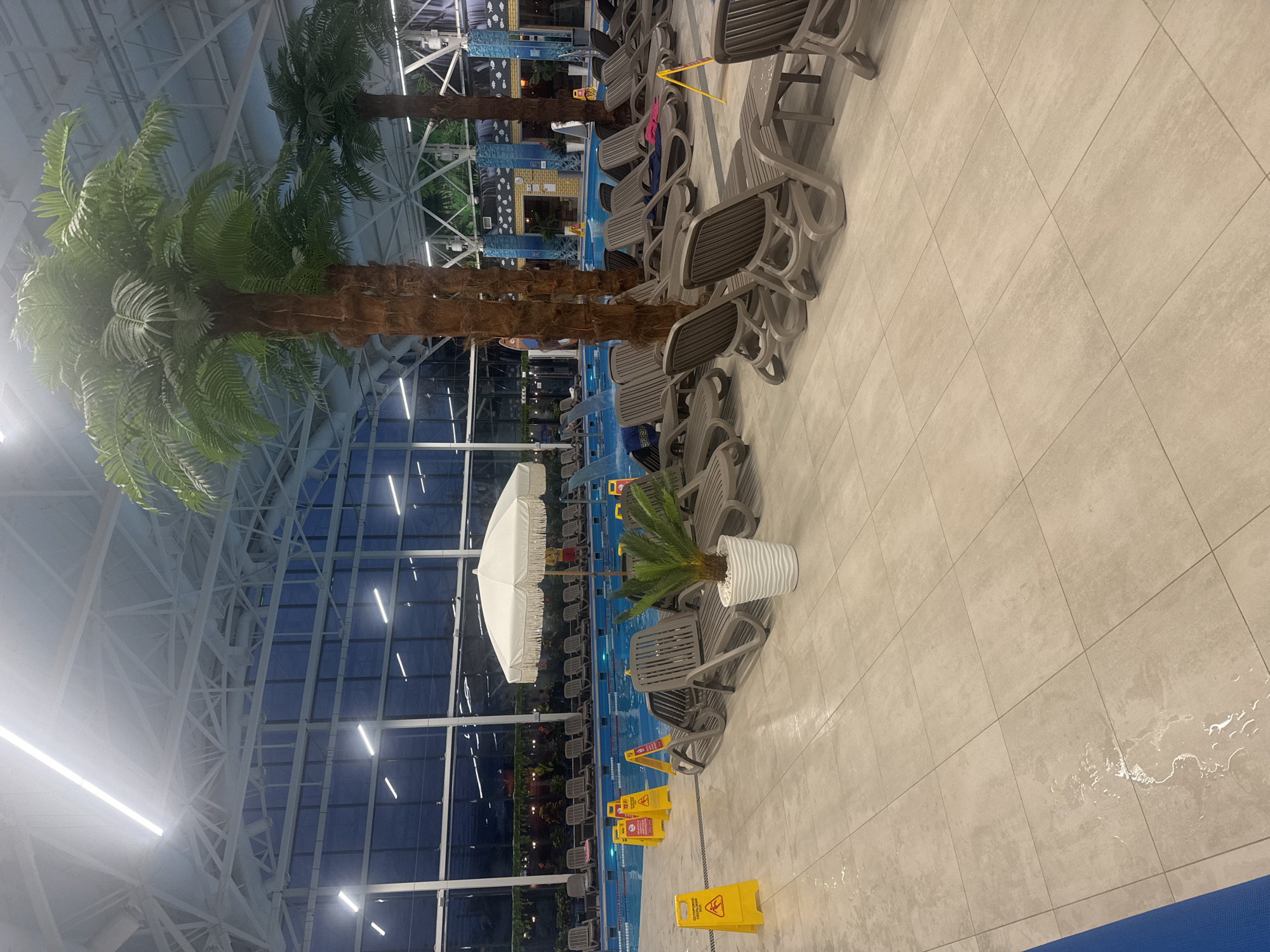 The image shows an indoor water park in Ulyanovsk, Russia. There are rows of grey lounge chairs lined up next to a blue pool. Palm trees and other greenery are scattered throughout the area. There is a white umbrella. The floor is tiled and there are yellow "Caution Wet Floor" signs present. The building has a large glass ceiling with a grid-like support structure. There are no people visible in the shot.