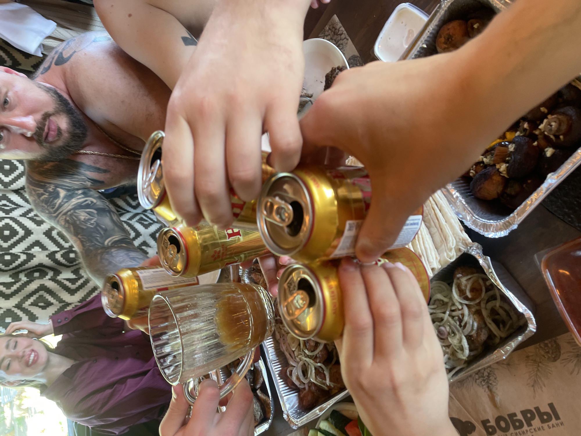 This media file captures a lively social gathering, likely outdoors or in a semi-enclosed recreational area, possibly a "Bobry Siberian Baths" complex in Дрокино, Russia, as indicated by visible text.
The central activity involves multiple hands clinking golden beer cans together and pouring their contents into a large, ribbed glass beer mug, which is partially filled with a yellow liquid. This suggests a shared toast or a communal drinking moment among friends.
Around this activity, a spread of food is visible, laid out in aluminium foil containers on a table. These dishes include dark, roasted items (possibly mushrooms or other grilled foods) with a white dressing, as well as meat pieces (likely shashlik or grilled kebab) generously topped with sliced white onions. Another container holds fresh, sliced vegetables such as cucumbers and tomatoes, and a smaller rectangular container contains a creamy white sauce or dip. Stacks of flatbread (lavash) are also present.
Two individuals are partially visible. In the upper left, a shirtless man with a beard, a gold chain, and extensive tattoos covering his arms and chest is seen looking towards the camera, resting on a black and white geometric patterned fabric. To the bottom left, a smiling woman in a long-sleeved purple shirt also looks towards the camera, conveying a cheerful mood.
The scene is well-lit by natural daylight, suggesting a daytime event. The overall atmosphere appears relaxed and jovial, typical of a casual meal and drinks with companions. Visible text on a brown surface confirms the name "БОБРЫ" (Bobry, meaning Beavers) and "СИБИРСКИЕ БАНИ" (Sibirskie Bani, meaning Siberian Baths), providing a specific location cue.