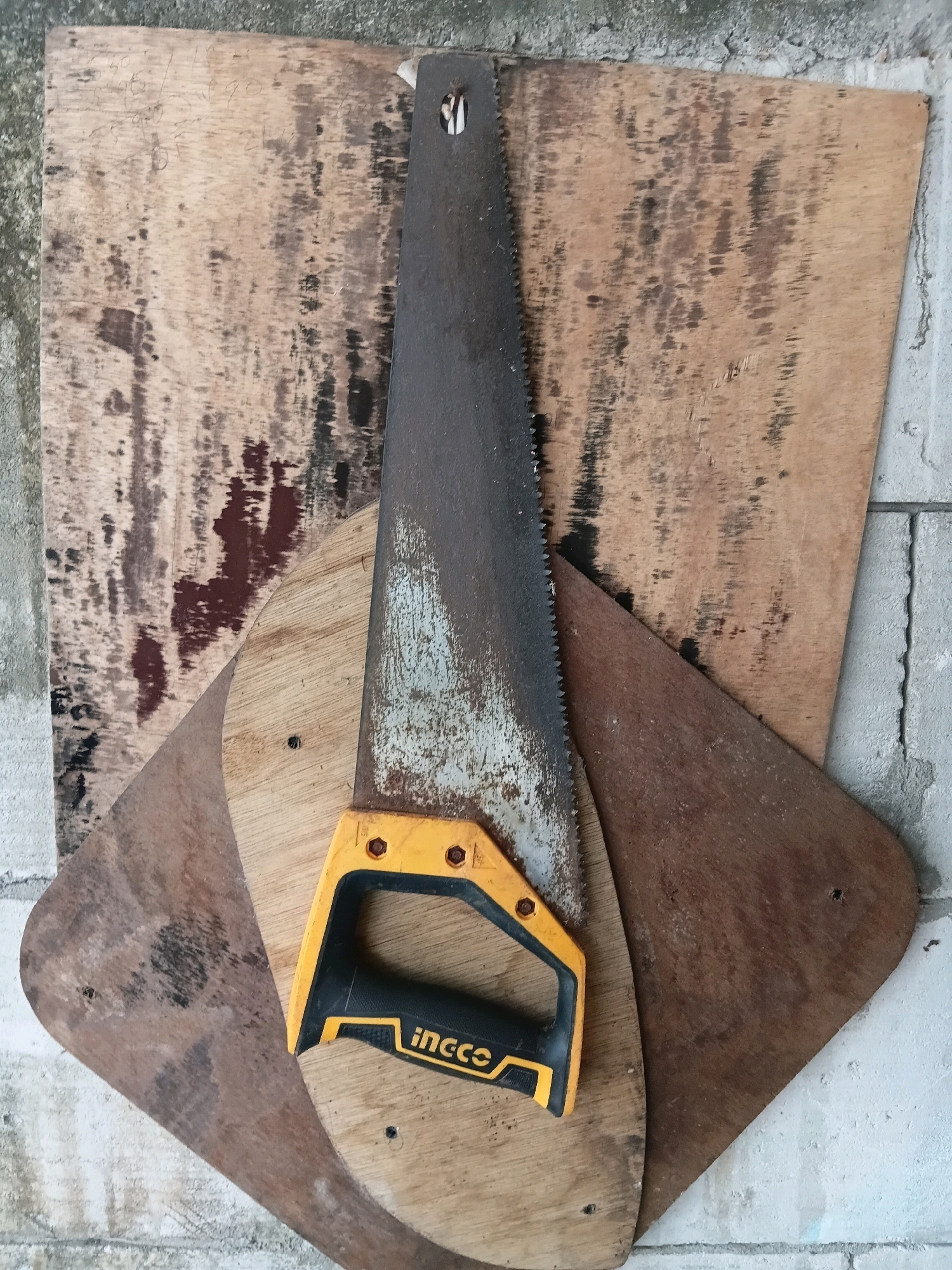 A hand saw with a yellow and black handle is placed on top of several pieces of plywood. The plywood pieces vary in size and shape and are arranged in a layered fashion. The background appears to be a wall made of concrete blocks. The overall scene suggests a workshop or construction area. The image was taken in the city of Cikal, Indonesia.