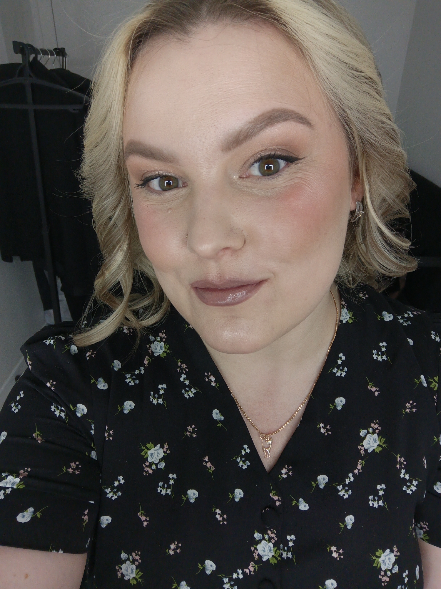 A fair-skinned woman with blonde, wavy hair poses for a selfie. She has brown eyes, and wears makeup including eyebrow pencil, eyeshadow, mascara, blush, and lip gloss. She wears a black floral print dress with a gold necklace featuring a cat pendant. The location is Unknown, Unknown. The woman appears to be indoors. A black garment is visible on a rack behind her.