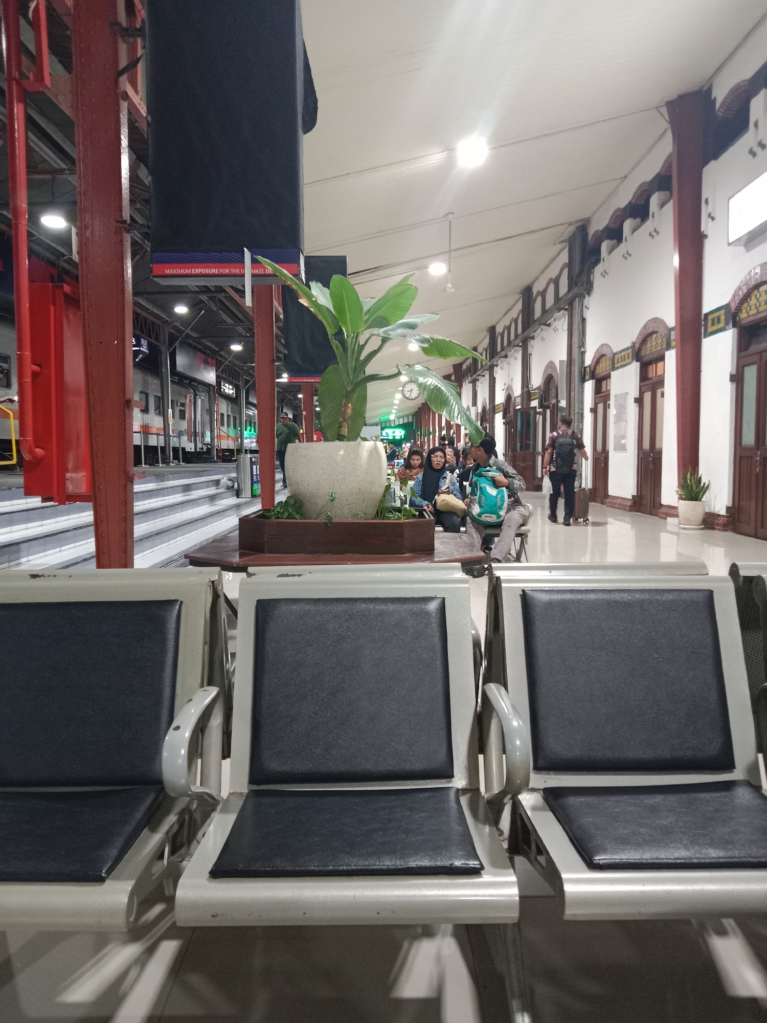 This is a low-angle, eye-level, medium shot of a waiting area at a train station in Semarang, Indonesia. The foreground features three empty, gray, metal chairs with black leather seats. Beyond the chairs, a wide walkway is visible, lined with white walls and arched doorways. A plant in a large pot stands to the left of the walkway, near a group of people sitting on benches. To the left of the plant, a train is visible at the platform, with stairs leading up to it. The train cars are white with orange accents. The lighting suggests it is either early morning or late evening. People are seen in the background, some sitting and waiting, while others are standing and walking. One person is seen pulling a suitcase, and another is carrying a backpack. The overall impression is that of a functional and somewhat busy train station.
