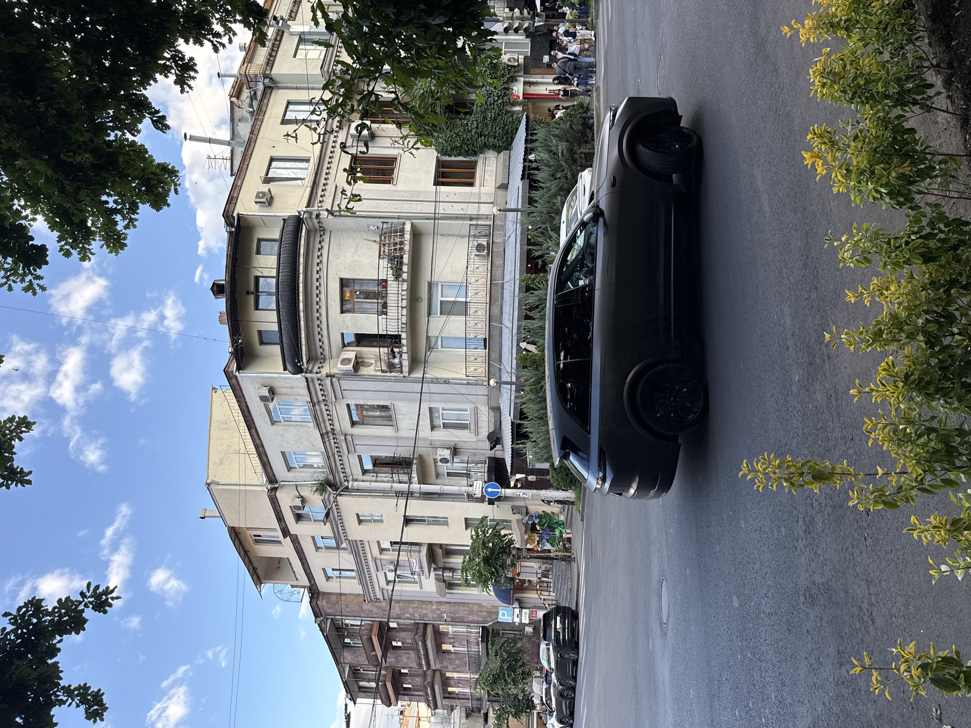 An outdoor, street-level photograph captures a sunny daytime scene in what appears to be a city, identified as Yerevan, Armenia. The image is rotated 90 degrees counter-clockwise.
The foreground features an asphalt road where a dark-colored SUV, possibly black or dark grey, is prominently parked on the right side. Its front wheel and the sleek profile of the vehicle are clearly visible. To the far right, a bush with vibrant green and yellowish leaves borders the street. The top left corner and bottom left edge are framed by dense green tree foliage.
Lining the street on the left are several multi-story buildings, characteristic of urban architecture. These structures display a mix of light-colored facades, some with intricate balconies featuring ornate railings. Windows of various sizes are visible, and several air conditioning units are affixed to the building exteriors. Some buildings appear older, possibly stone-clad in darker tones, while others are lighter beige or cream. Satellite dishes and chimneys are discernible on rooftops.
Further down the street, a group of people are gathered on a sidewalk, appearing small in the frame. They seem to be standing or waiting near what might be a cafe or shop entrance. Street furniture, including a blue circular sign with a white left-pointing arrow and a partially visible blue "P" parking sign, are present.
The sky above is a bright blue with scattered white, puffy clouds, indicating clear and pleasant weather. The overall impression is that of an ordinary, calm urban street scene during daylight hours.