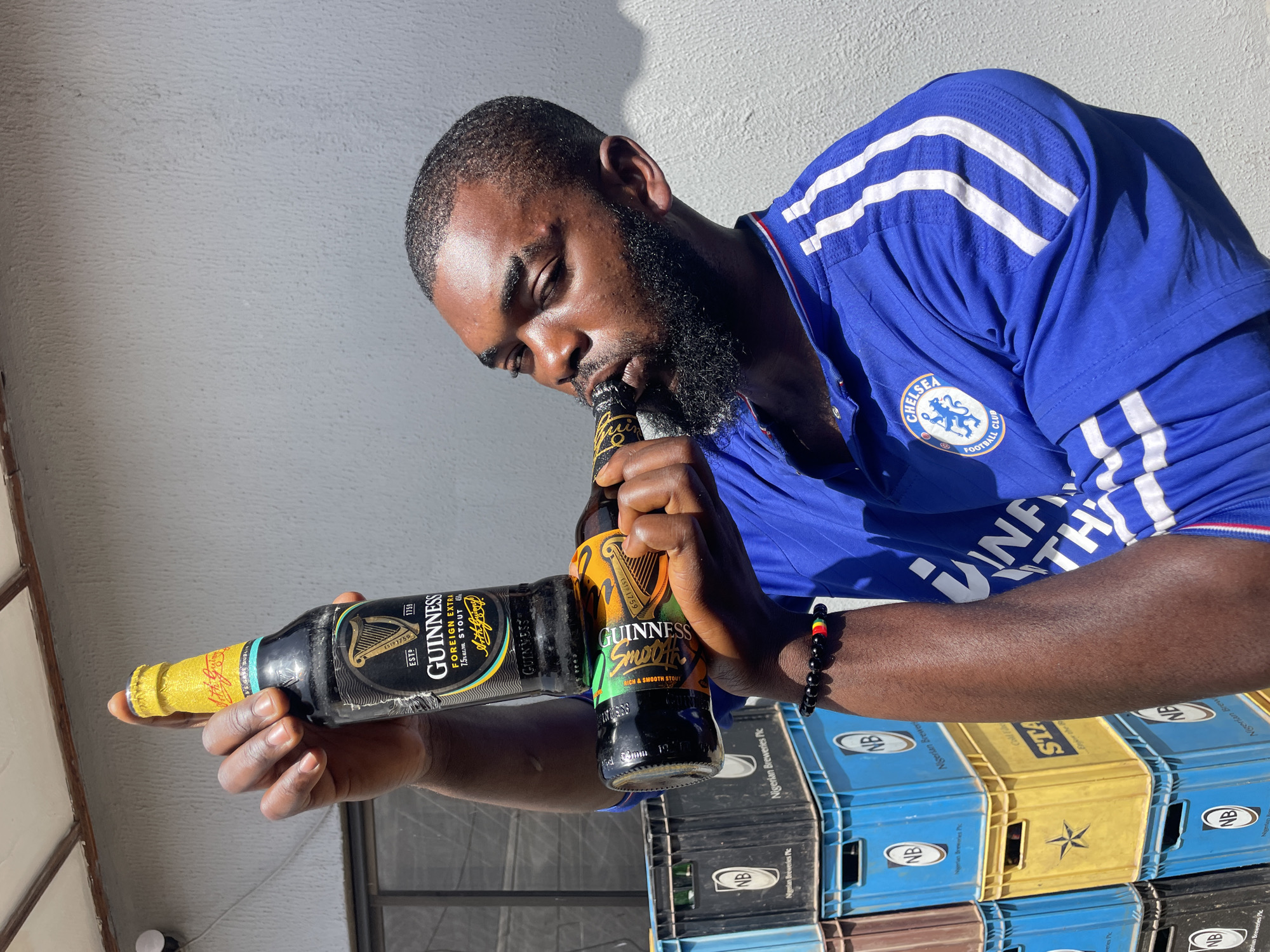 A man is holding two bottles of Guinness beer. He is wearing a blue Chelsea Football Club jersey. One bottle is Guinness Foreign Extra Stout, and the other is Guinness Smooth Rich & Smooth Stout. He is looking down at the bottles, with one bottle near his mouth. Behind him are stacks of plastic crates, some blue and some yellow, with "NB Nigerian Breweries Plc" visible on them. The scene appears to be outdoors, with a rough, textured wall in the background and sunlight casting shadows. The time of day seems to be daytime, likely afternoon, due to the strong shadows. The man has a beard and short hair.