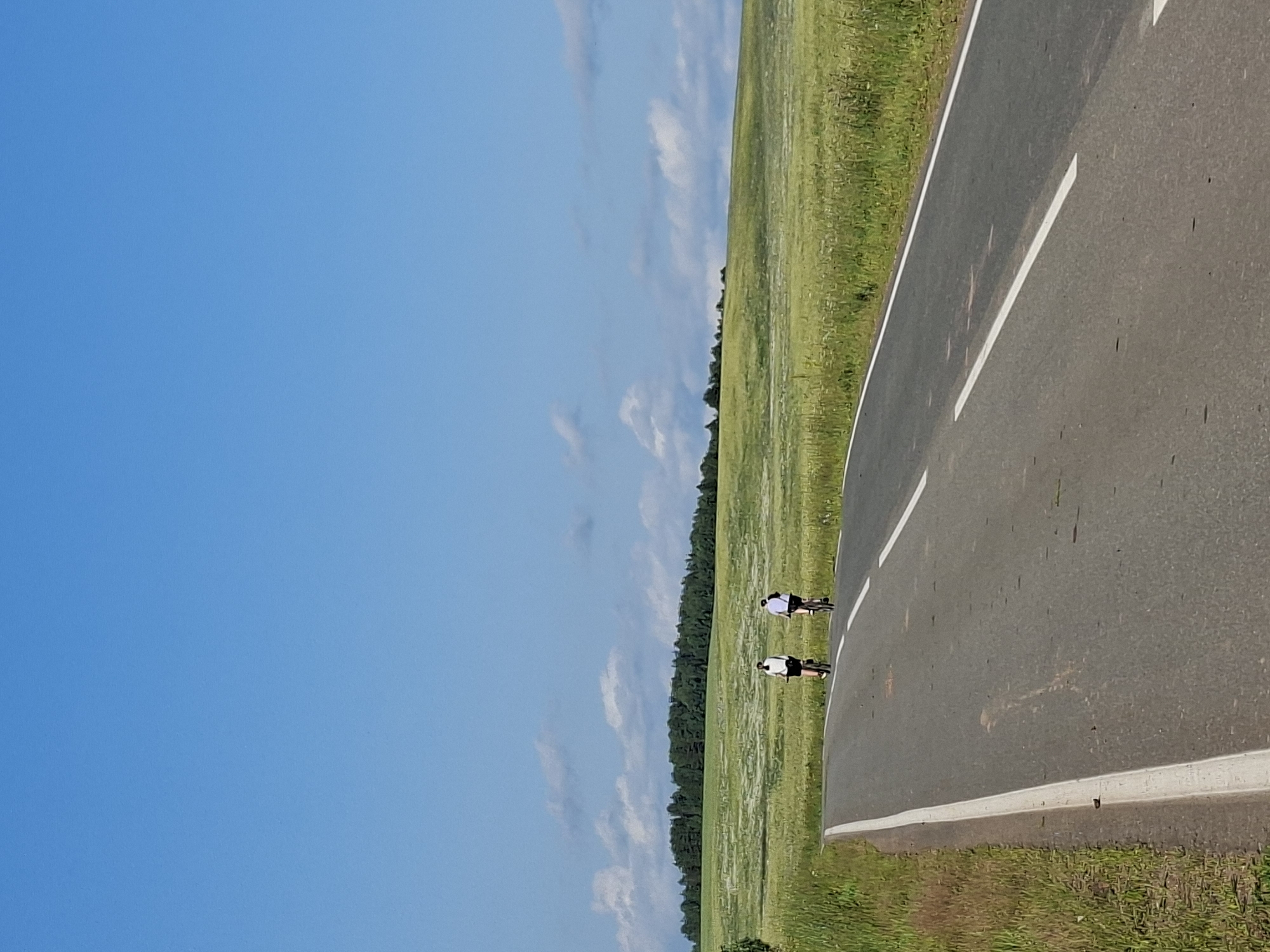 Two individuals are cycling on a paved road bordered by a vibrant green field under a clear blue sky with scattered white clouds. The road has distinct white lane markings. The field is lush, with some areas appearing lighter, possibly indicating wildflowers or a different type of grass. A line of trees forms the distant boundary of the field. The perspective suggests the photo was taken at eye level, with the road curving away into the distance. The lighting and clear sky indicate it is daytime, likely a sunny day. The overall scene evokes a sense of outdoor activity and peacefulness. The location is identified as Chumna, Russia.