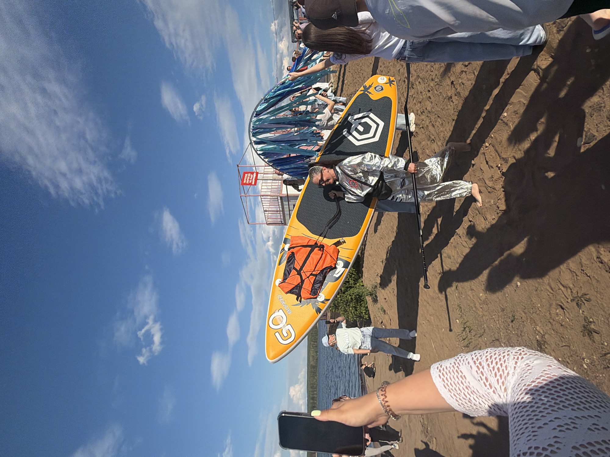 This image depicts an outdoor gathering on a sandy shore, likely near a body of water given the presence of a lake or river in the background and a clear blue sky with scattered clouds. Several individuals are present, with one person prominently featured in the foreground carrying a large orange paddleboard. This individual is wearing a shiny, silver suit and appears to be barefoot. Other people are visible in the mid-ground and background, some holding phones, suggesting they might be taking pictures or videos. A woman in the mid-ground is wearing a light-colored t-shirt and jeans, holding a phone and a paddleboard. In the background, a colorful, arch-like structure draped with blue and white fabric is visible, along with a red sign with text in Russian. The overall atmosphere appears to be casual and festive. Based on the provided context, this scene is located in the city of Chaikovsky, Russia.