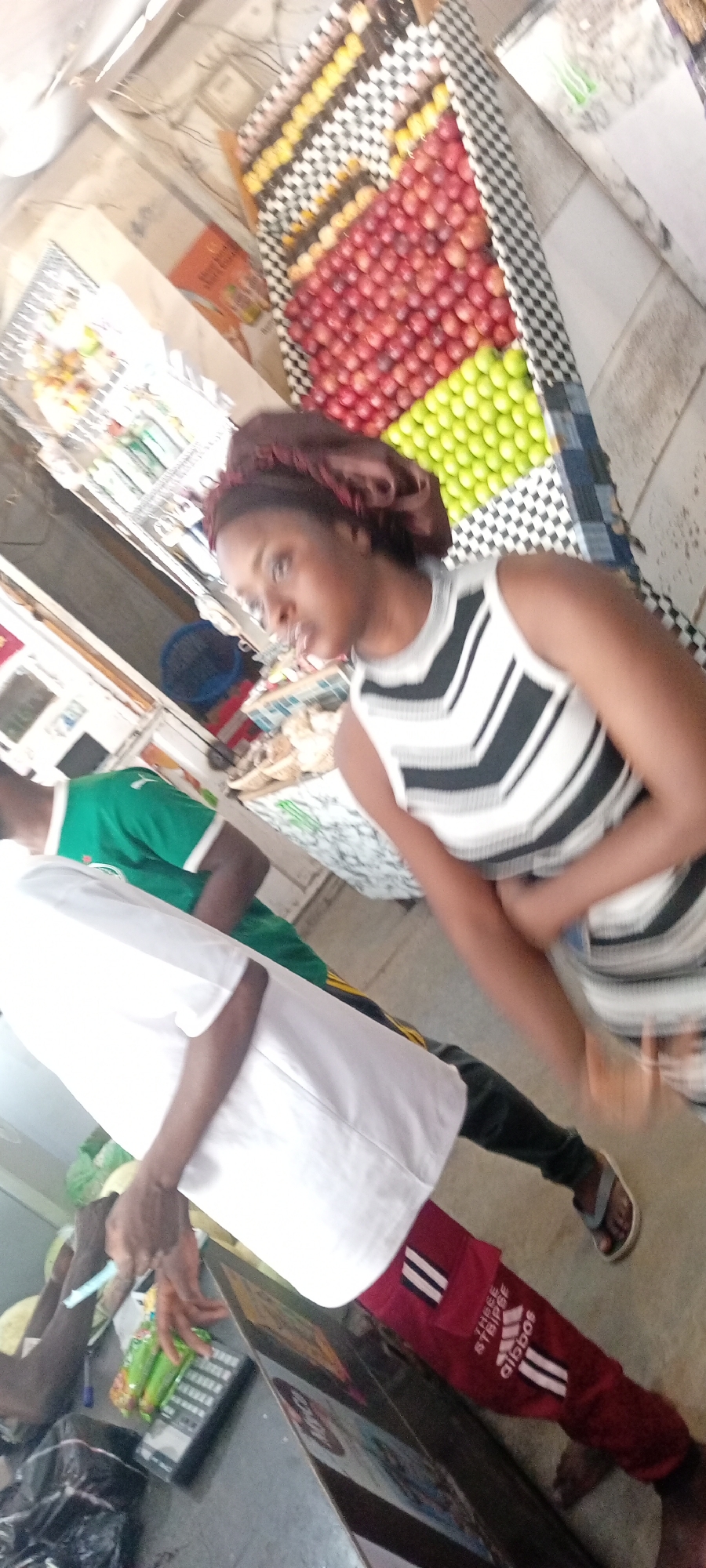 Here's a description of the image:
**Location:** Aviation Quaters, Nigeria
**Scene:** An indoor commercial space, likely a market or a store, given the presence of produce and a point-of-sale system.
**People:**
* A young woman with a head wrap, wearing a striped top, is in the foreground. She appears to be a customer.
* A person wearing a white top, green shirt, and red pants with Adidas stripes appears to be working at a counter or till.
**Objects:**
* Behind the woman is a display of fruit (apples, limes) organized in neat rows.
* A Point-of-sale (POS) system is on the counter.
* There are other products and containers visible in the background, on shelves.
**Actions/Interactions:**
* The person wearing the white top appears to be using the POS machine, possibly processing a transaction.
* The woman in the striped top is interacting with the counter area.
**Environment:**
* The lighting appears to be a mix of natural and artificial.
* The store has a tiled floor.