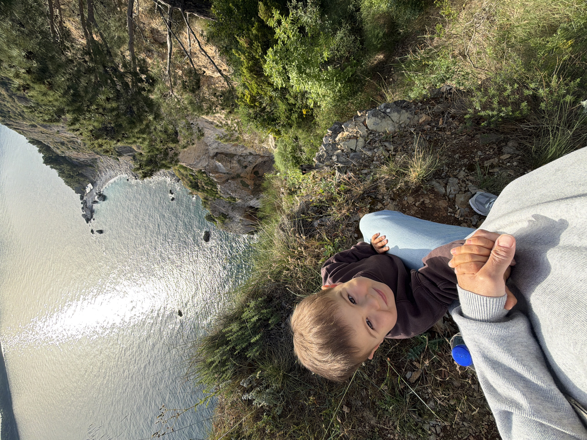 The image depicts a person and a young child near a cliff overlooking the Adriatic Sea. The location is suggested to be near Budva, Montenegro, based on the coastal landscape. The child, wearing a brown sweater, is looking up at the camera. An adult, wearing jeans and a grey sweater, is visible from the torso upwards, and their hands are holding the child's hands. The cliff is grassy with shrubs and a few trees. The sea reflects sunlight. A water bottle is visible on the ground near the person.