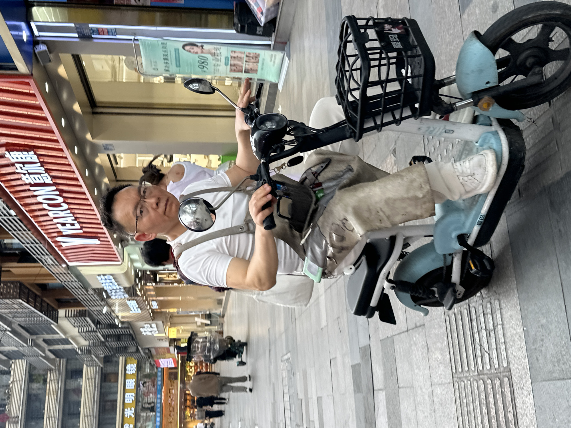 A medium shot captures a man on a light blue e-bike, looking toward the viewer in Jiangjin, China. He wears glasses and a white shirt with a shoulder bag. He sits with his hand on the handlebar. The e-bike features a black wire basket on the front and a small seat. In the background is a paved pedestrian street flanked by shops and stores. Signs in Chinese characters are visible above the shops. Additional pedestrians can be seen walking in the background. The image is slightly rotated.