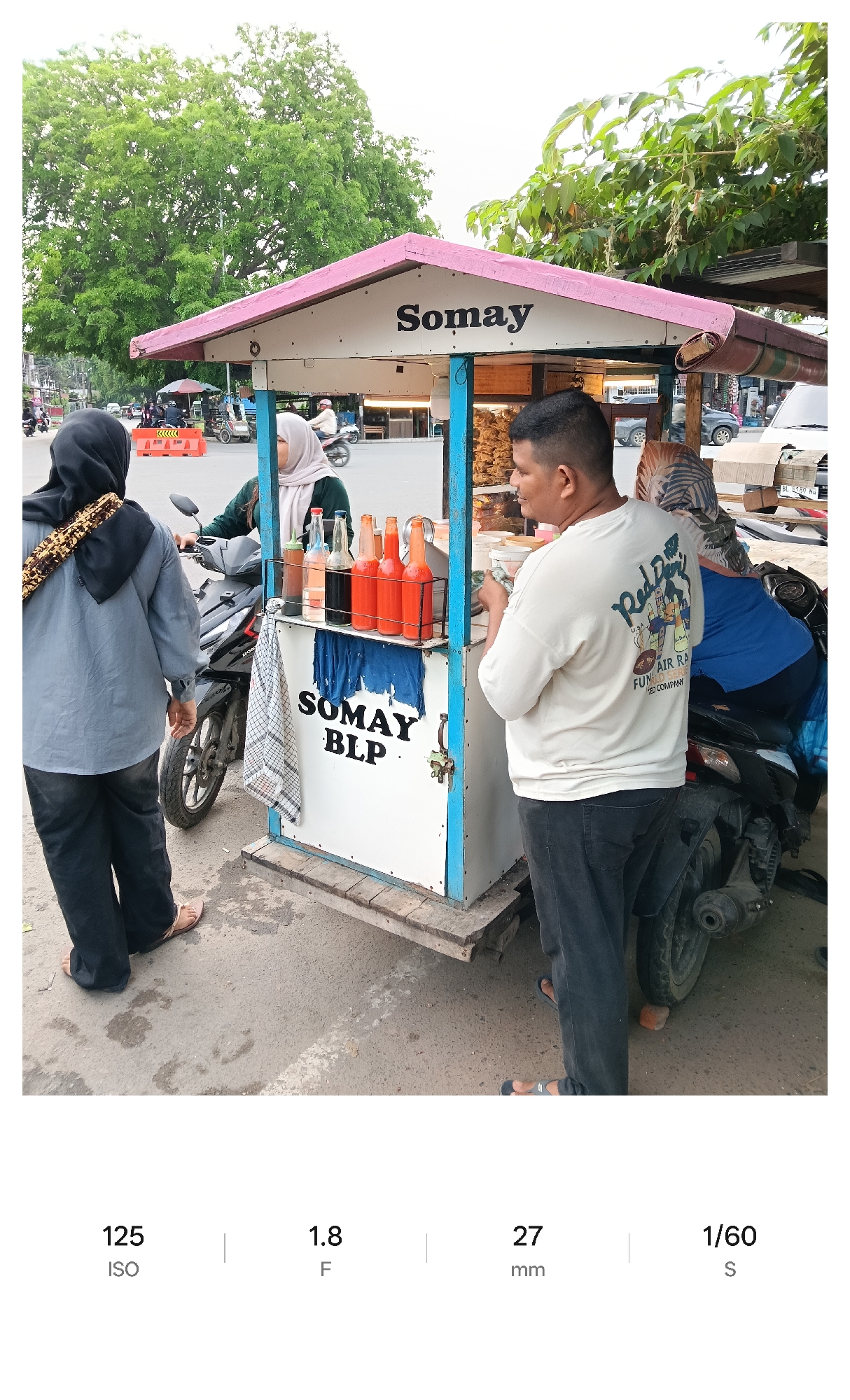 Captured outdoors on a street in Lhokseumawe, Indonesia, the image centers on a food vendor operating a small cart named "Somay BLP." The cart, painted with a white base, features a pink roof and blue trim. Clear glass bottles filled with liquids, some with a reddish-orange hue, stand on the cart's counter.
A man in a white t-shirt and dark pants appears to be purchasing food from the vendor. Another person, dressed in a gray shirt and black pants, stands nearby. A woman wearing a hijab and a green shirt is visible on a motorcycle. Another woman, who also has a hijab, is on a motorcycle behind the man.
In the background, trees and buildings are visible, indicating an urban setting. A road with vehicular traffic adds to the scene's vibrancy. The time of day is likely daytime, given the ample natural light. The presence of the food cart and motorcycles indicates a bustling local scene.