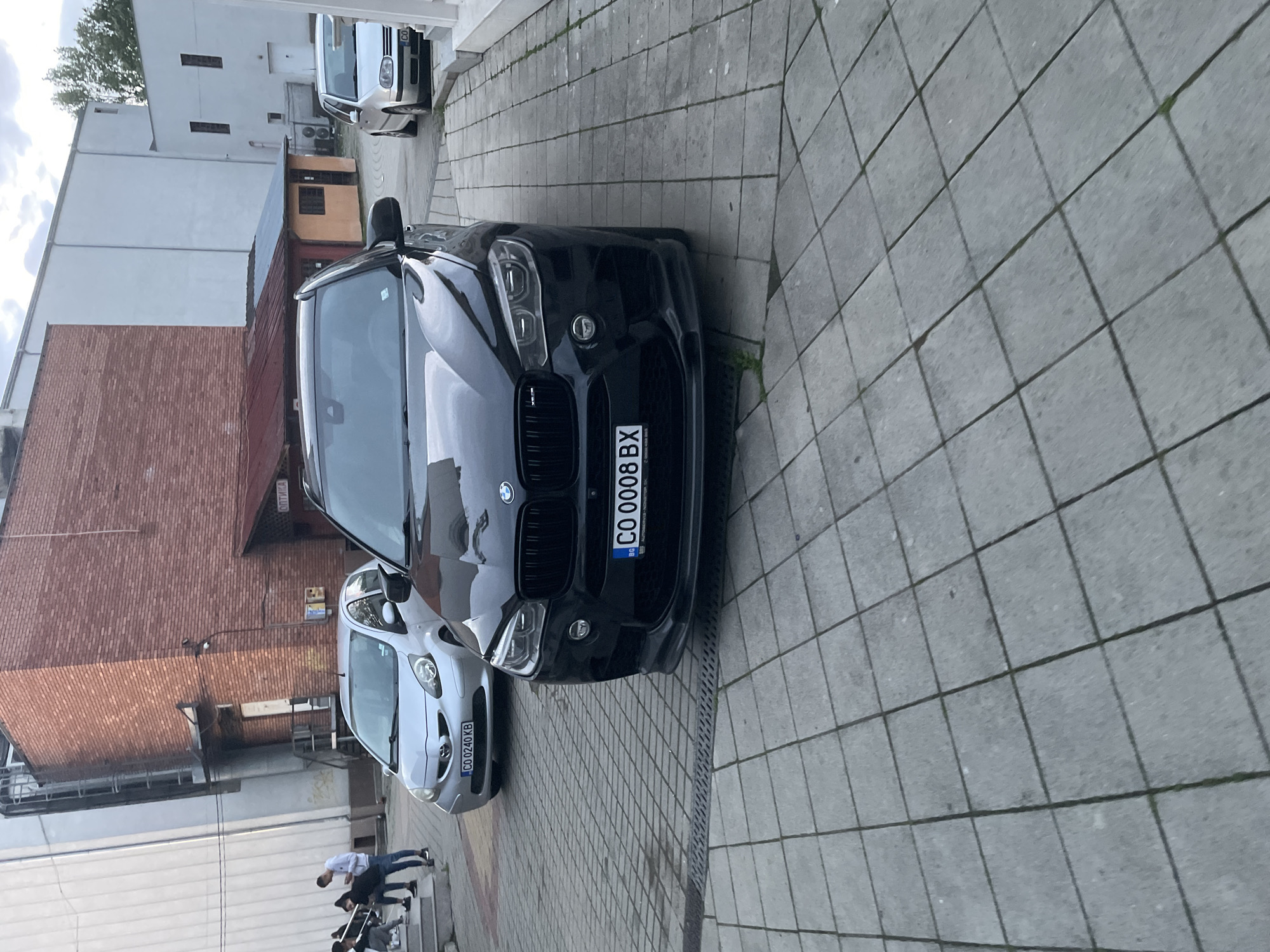 This image captures a street scene in Ihtiman, Bulgaria, during what appears to be an overcast day, possibly late afternoon or early evening. The main subjects are two cars parked on a paved area, flanked by buildings.
In the foreground, a black BMW SUV (likely an X5) is prominently parked. Its front license plate, clearly visible, reads "CO 0008 BX" with the blue Euro band indicating "BG" for Bulgaria. Behind the BMW, a silver Toyota Aygo is parked, displaying the license plate "CO 0240 KB," also with the "BG" country code. Further in the background, a white Volkswagen Golf Mk4 is partially visible, parked near a white building; its license plate also begins with "CO." The cars are situated on a street or alleyway paved with large, grey, rectangular tiles. Some grass or weeds are growing through the cracks in the pavement, indicating a less trafficked or older area.
On the left side of the frame, several buildings are visible. There's a tall, plain white building, and behind the Toyota, a brick-faced building with a prominent sign that partially reads "ОПТИКА" (OPTICA) in Cyrillic, suggesting an optician's office or shop. Between these two buildings, a smaller, reddish-brown structure, possibly a shed or kiosk, can be seen.
In the lower left background, several people are present. One man in a white shirt and dark pants is standing, while others are seated or partially visible, suggesting casual activity in the area. The overall impression is a quiet urban street scene.