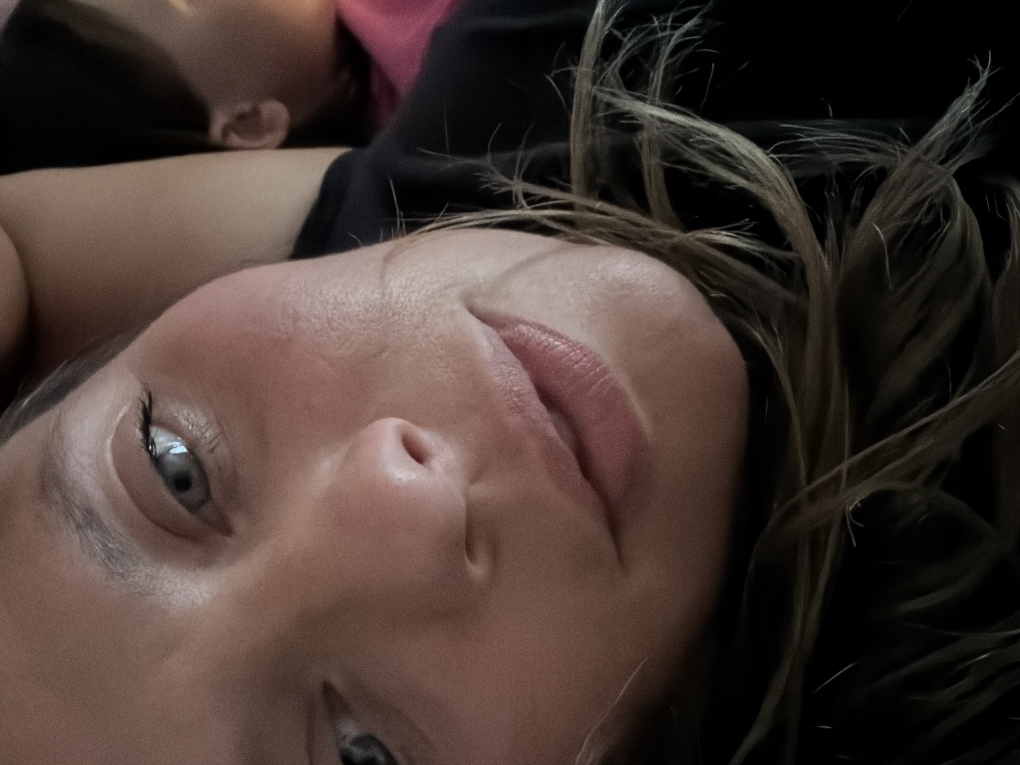This media file is a close-up, rotated image of a person's face. The person appears to be lying down, with their head tilted back. Their eyes are partially open, and their lips are parted slightly. The lighting suggests it might be indoors or in a dimly lit environment. The background is dark, with strands of hair obscuring some of the details. There are no visible objects or text in the image. The context provided places this scene in Kamianka-Buzka, Ukraine, but there are no visual cues within the image to confirm this location. The overall mood is intimate and possibly relaxed, given the recumbent pose.