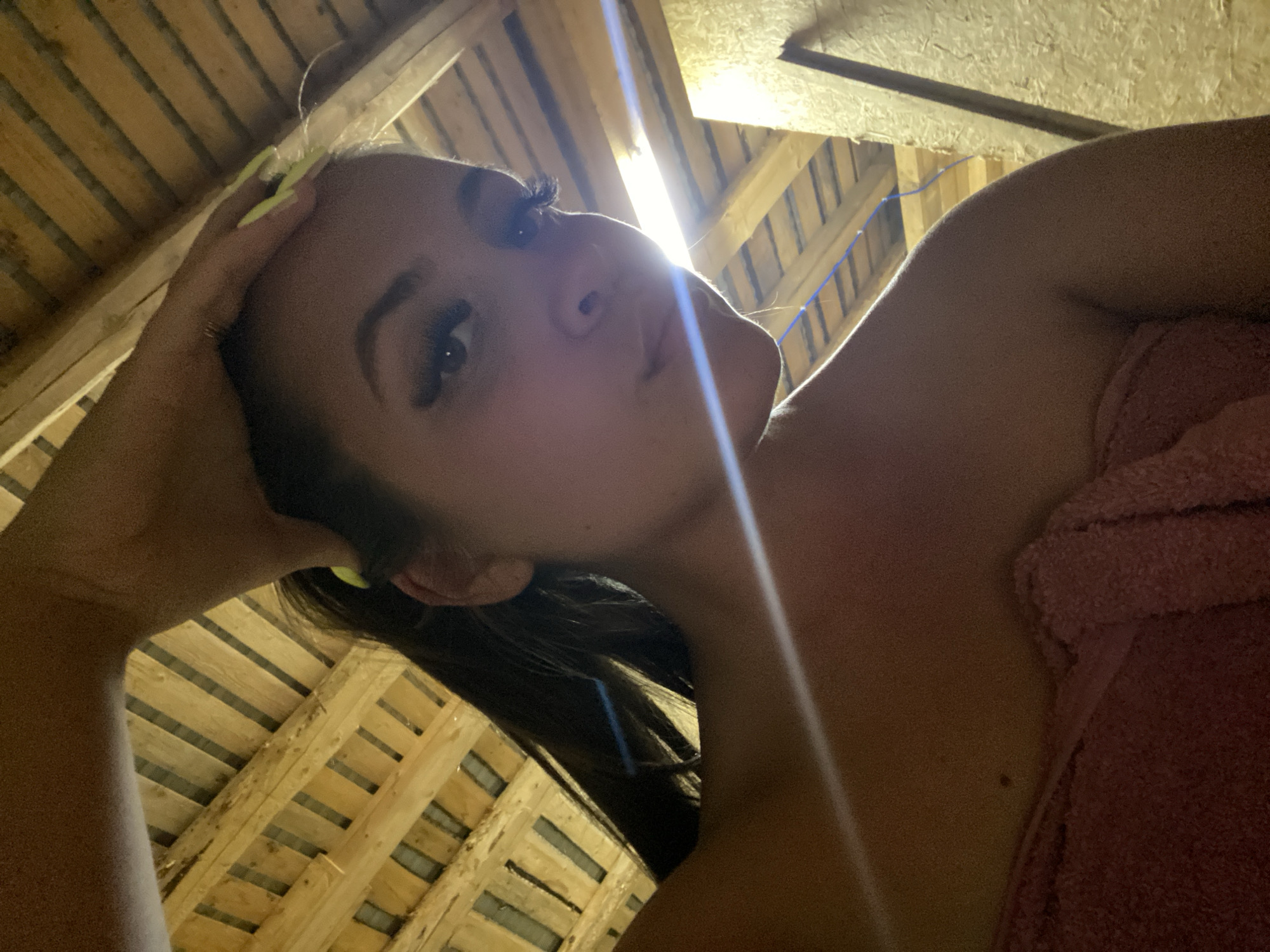 A young woman with long dark hair pulled back is captured in a low-angle, selfie-style shot. She is wearing a pink towel wrapped around her upper body and is holding her right hand to her forehead. Her makeup includes dramatic false eyelashes. The background consists of wooden beams and planks, suggesting an attic or unfinished structure. A strong light source, possibly sunlight, creates a lens flare across the image. The setting appears to be within the village of Zhigalki, Russia.