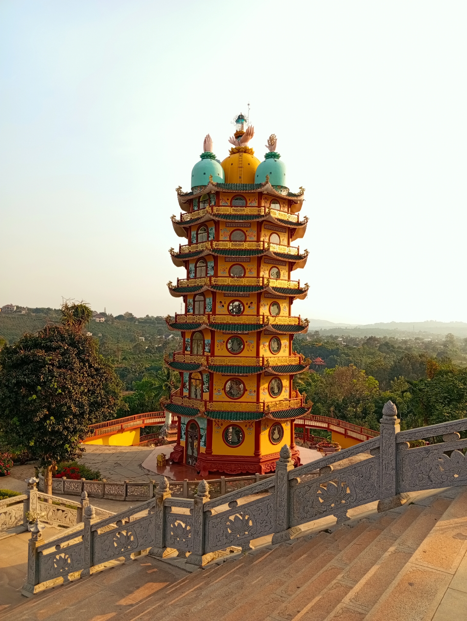 The image shows a tall, colorful pagoda with multiple tiers, likely a Buddhist temple. The structure is predominantly yellow and red, with decorative green and gold accents. Each tier features windows, and the topmost tier is crowned with yellow and green domes. The pagoda is situated on a raised platform, surrounded by trees and greenery, giving the impression that it is located on a hill or elevated terrain. In the foreground, there are stone stairs and ornate railings leading up to the pagoda, suggesting a path for visitors. The background consists of a hazy sky and distant hills or mountains. The picture appears to be taken during the day, with soft, natural lighting. Based on the information provided, it is located in the city of Phú An, Vietnam.