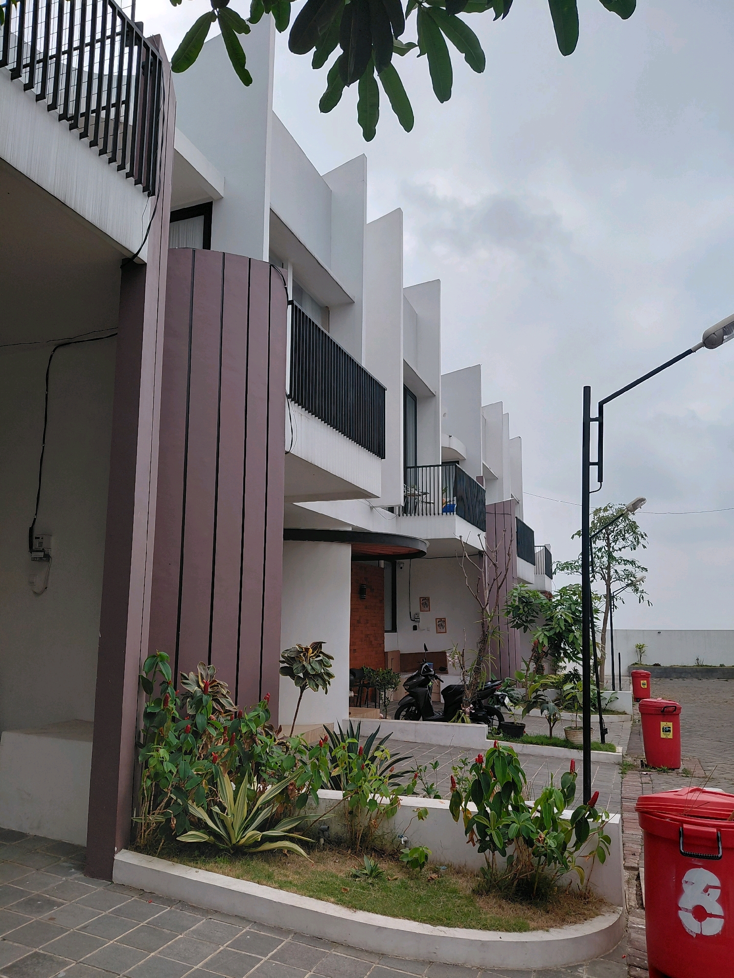 In Batu, Indonesia, a row of modern, multi-story townhouses with white facades and dark railings lines a paved walkway. Lush green plants and red-flowered bushes are neatly arranged in planter boxes along the front of the buildings. A tall streetlamp stands on the right side of the image, casting light on the scene, and two red trash bins are positioned near the street. The sky is overcast, with a few clouds visible. A portion of a tree with large green leaves is visible in the upper left corner of the image.