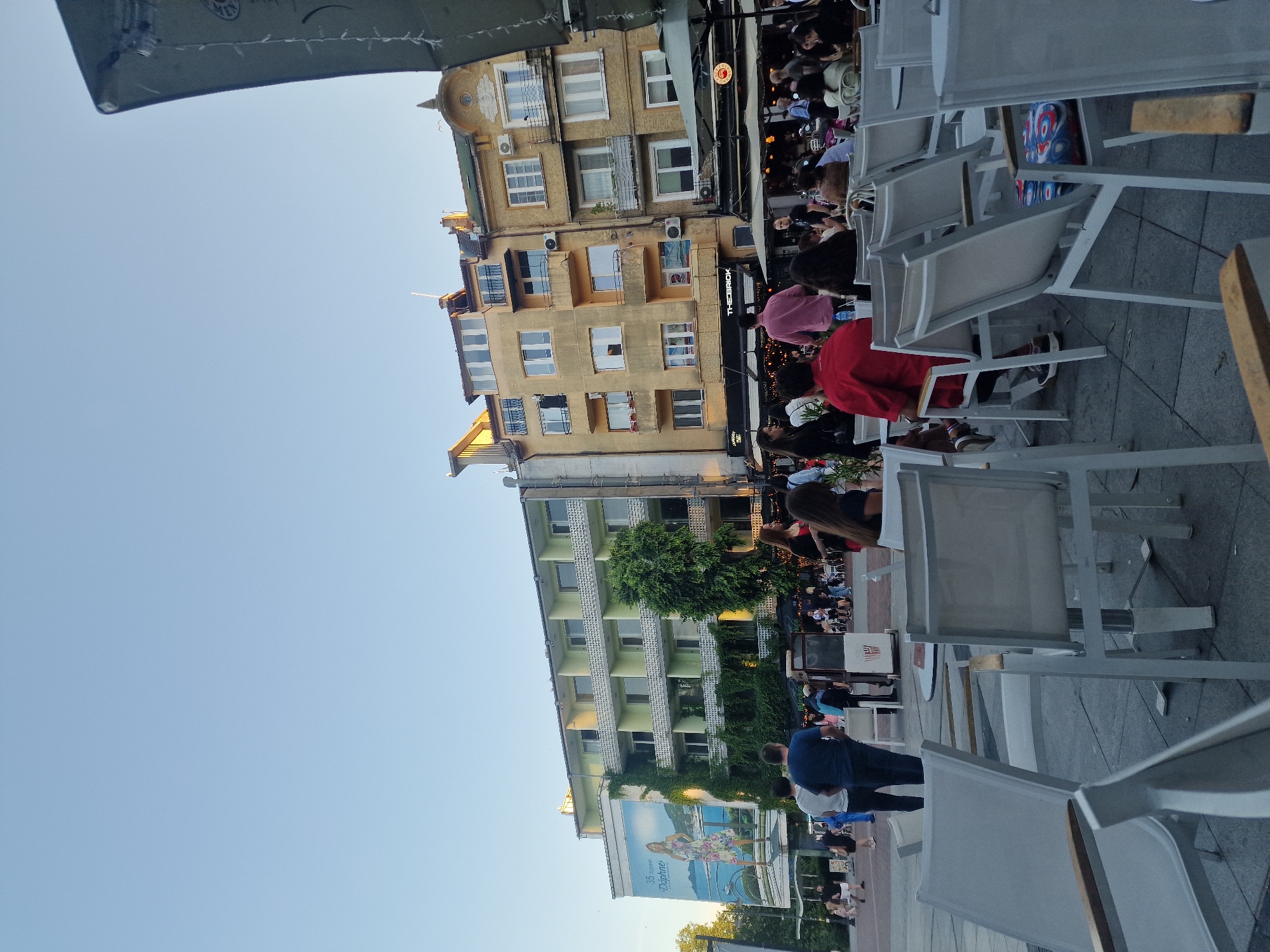 The image depicts an outdoor scene in a city square, likely a pedestrian zone with cafes. In the foreground, white metal outdoor chairs and tables are arranged on a paved surface. A few people are seated at the tables in the midground, and others are walking in the background. The backdrop consists of several multi-story buildings. The building on the left is light green with balconies and a large mural featuring a woman and a bicycle. Adjacent to it are two yellow buildings with numerous windows and balconies. Several cafes with awnings and outdoor seating are visible in front of the yellow buildings. The sky is clear and blue, suggesting daytime. The overall atmosphere appears to be casual and social, with people enjoying the outdoor space.
