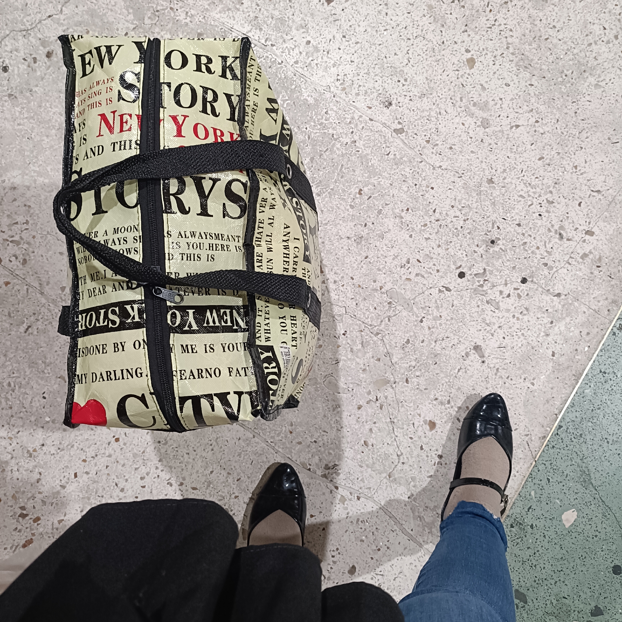 The image shows a New York Story tote bag with black straps and a zipper. The bag is made of a white material with black text and red accents, including a heart shape. The text on the bag appears to be in a newspaper print style, with phrases like "New York Story" and other writings. The bag is positioned on a speckled gray floor, possibly terrazzo, with a light gray grout. To the right of the bag, a pair of black, pointed-toe heels with a strap are visible, worn with tan stockings and blue jeans. The floor also has some subtle cracks. The overall lighting suggests an indoor environment. The image is cropped to focus on the bag and the lower half of a person. There are no discernible landmarks or clues to suggest a specific location within Semarang, Indonesia.