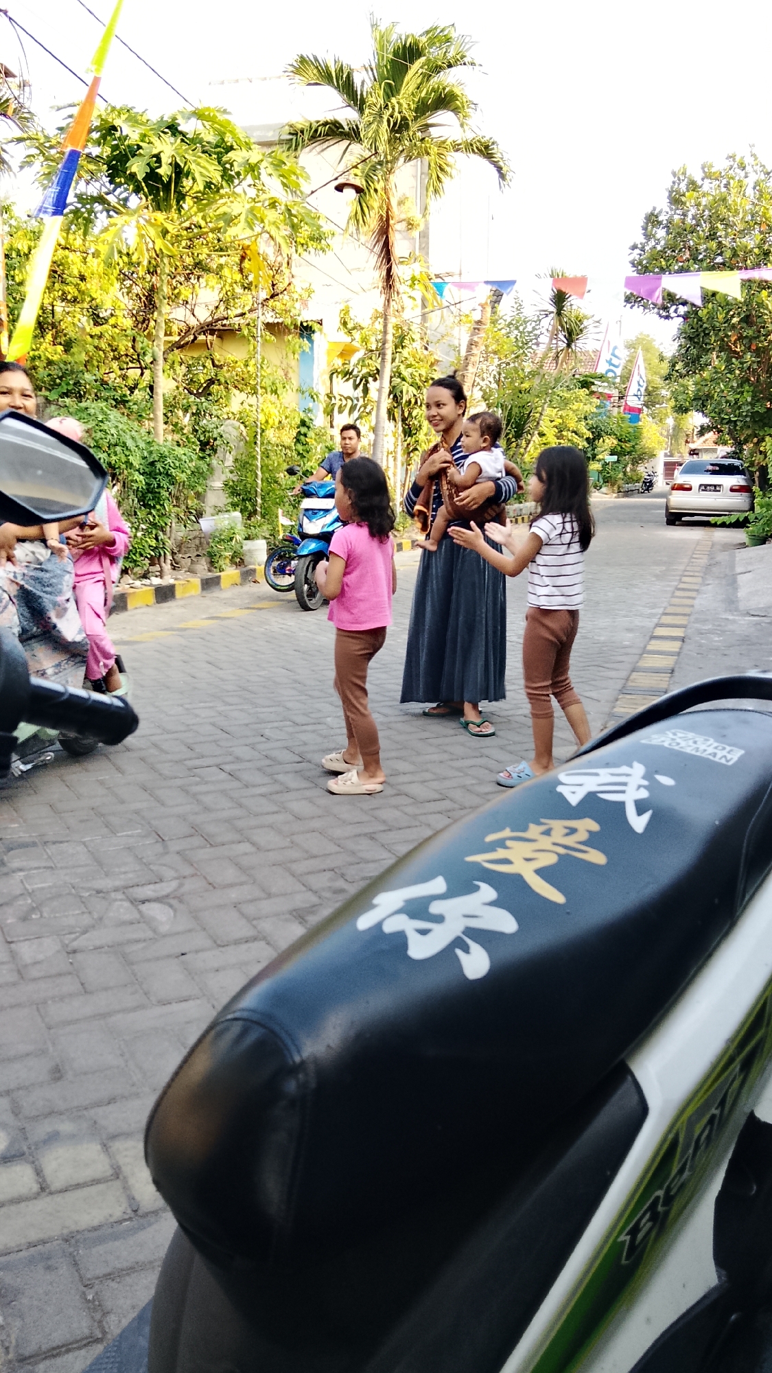 In Banjarsari, Indonesia, a woman stands holding a baby, with two young girls nearby on a paved street lined with trees and buildings. One girl in a pink shirt and brown leggings is facing away from the camera, while another in a striped shirt and brown leggings is facing the woman. A man on a blue motorcycle is visible in the background. The scene is decorated with colorful flags, suggesting a festive occasion. In the foreground, the seat of a motorcycle with Asian characters is prominent.