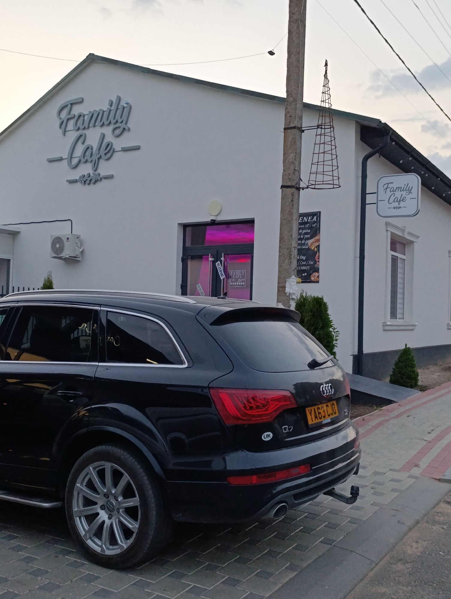 In the city of Cocieri, Moldova, a black Audi Q7 SUV is parked on a cobblestone street in front of a white building with the sign "Family Cafe" in large grey lettering. The cafe has a modern, minimalist design with a prominent window displaying bright pink lighting. A second, smaller "Family Cafe" sign is visible on the right side of the building. A utility pole stands to the left of the building, adorned with a conical wire structure. The sky is overcast, suggesting a late afternoon or early evening setting. The overall scene depicts a commercial establishment and its immediate surroundings.