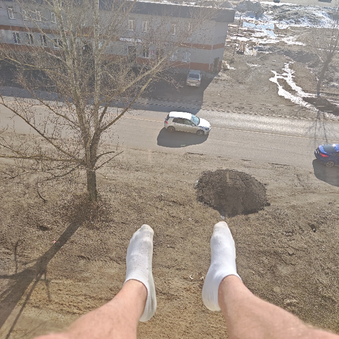 A high-angle perspective, taken from an elevated position in Rostov Veliky, Russia, captures an outdoor urban environment during daylight. In the lower foreground, the bare lower legs and feet of an individual, clad in white ankle socks, are extended into the scene. The immediate ground below consists of light brown, unpaved earth. A leafless deciduous tree stands on the left within this unpaved area, casting a shadow. To the right of the tree, a mound of dark soil is present. A paved road traverses the mid-ground, featuring a white 5-door hatchback traveling towards the right, casting a shadow, and a partial view of a dark blue SUV on the far right. Across the road, a multi-story building with a grey facade and orange horizontal accents is visible. Beyond this building, an expanse of disturbed, muddy ground with standing water and residual patches of snow or ice indicates a non-developed area. Bright ambient light with distinct shadows suggests sunny or partly cloudy conditions.
