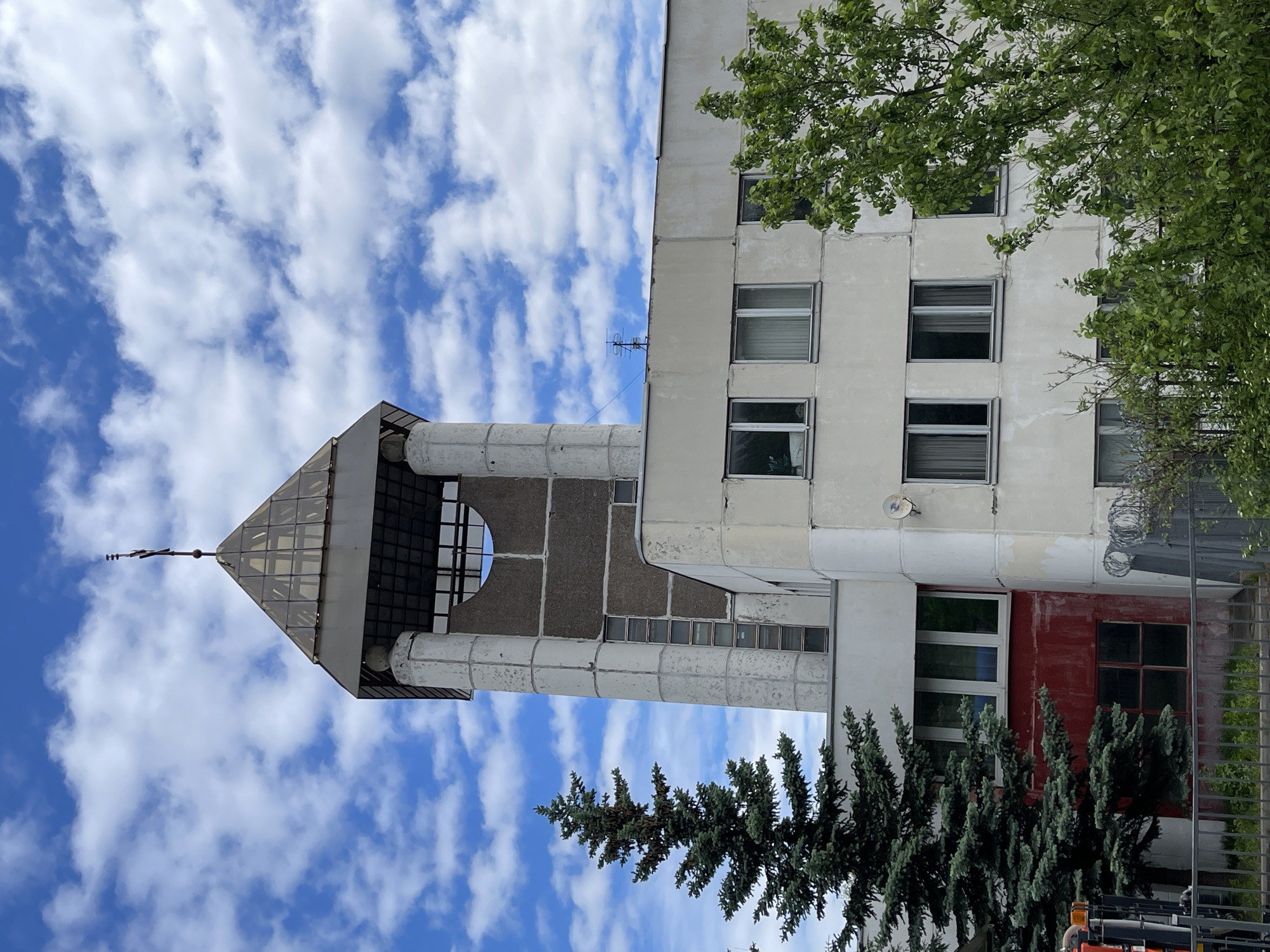 The image depicts a building with a distinctive tower and a modern architectural design against a sky filled with white clouds. The tower, featuring a geometric shape and a spire, appears to be an integral part of the building's structure. A section of the building is painted with a red panel, and there are several windows visible. Trees are interspersed in the foreground. Based on the architectural style and building characteristics, the location is likely in Moscow, Russia. The building is possibly a civic or institutional structure.
