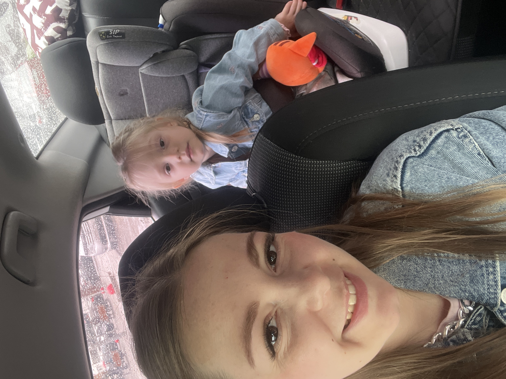 The image depicts a selfie taken from inside a vehicle, featuring an adult female in the foreground and a young child in the background. The location is an urban or suburban outdoor setting, visible through car windows, in the city of Unknown, Unknown, under rainy conditions.
The adult female, positioned in the front passenger seat, occupies the left and central foreground. She has light brown hair and a fair complexion, smiling directly at the camera. She wears a light-colored top under a denim jacket, adorned with a chunky silver-colored chain necklace. Her visible eyelashes appear enhanced, and her eyebrows are well-defined.
In the rear passenger seat, to the right and slightly behind the adult, a young child, appearing to be a girl between 2-4 years old, is secured in a gray child safety seat. The safety seat features "SIP" (Side Impact Protection) branding. The child has blonde hair, partially styled in a ponytail or pigtail on her right side, and wears a light blue denim jacket over a brown garment. She looks towards the camera with a neutral expression. An orange plush toy, possibly a fox, rests beside her in the car seat.
The car's interior features black seats with visible stitching and textured fabric on the front seat back. A gray grab handle is visible on the ceiling. Through the rear passenger window, numerous water droplets indicate rain. The exterior environment shows multiple parked vehicles, suggesting a parking lot or street. Some indistinct red objects are visible in the distance through the rain-streaked window. The overall lighting is diffuse, consistent with an overcast, rainy day.