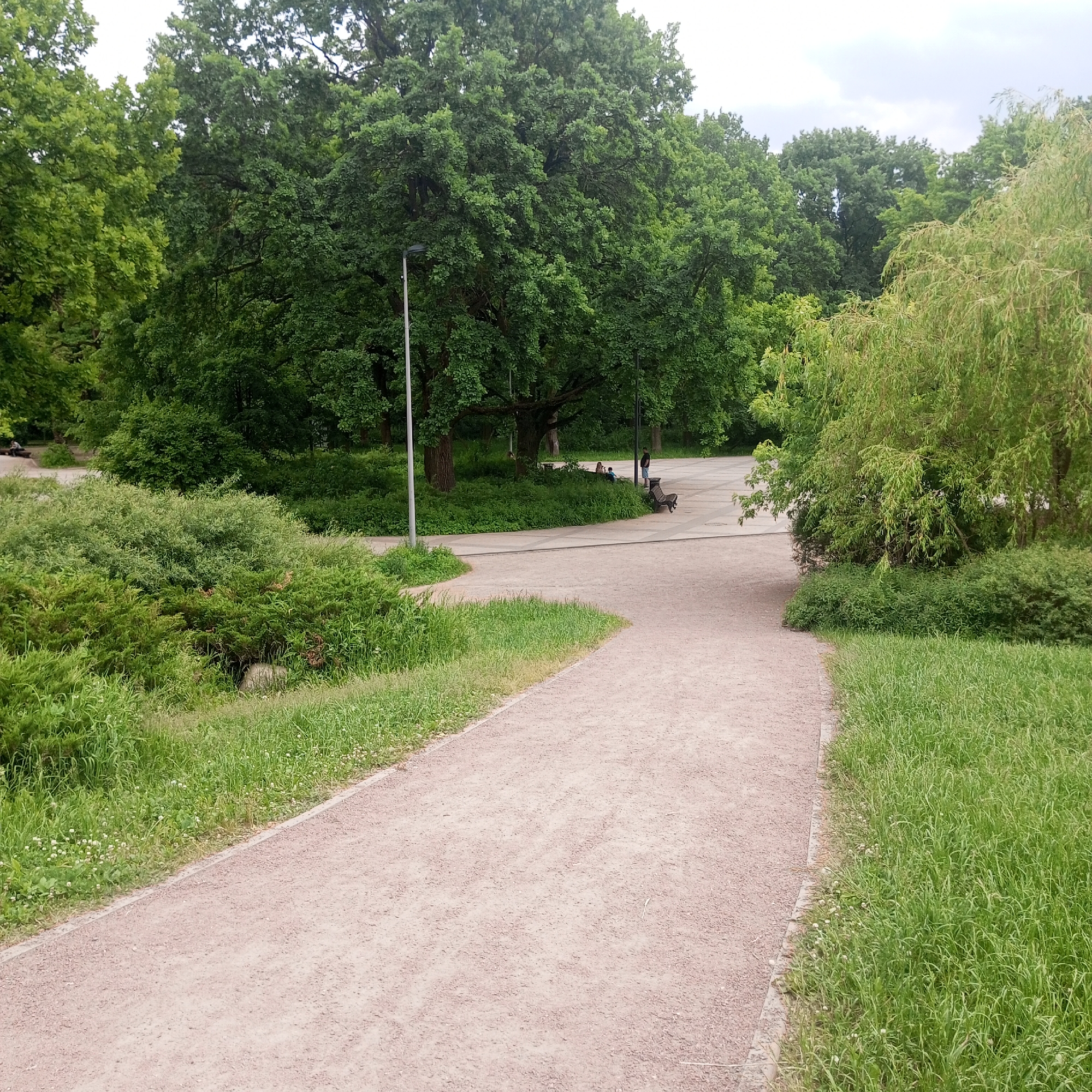 The image depicts a park path made of gravel, curving through a grassy area with trees. In the background, there are large trees and a paved area with a few people. One person is standing by a lamppost, while others are seated on a bench. The environment appears to be a public park in Moscow, Russia, likely during the daytime. The weather seems overcast.