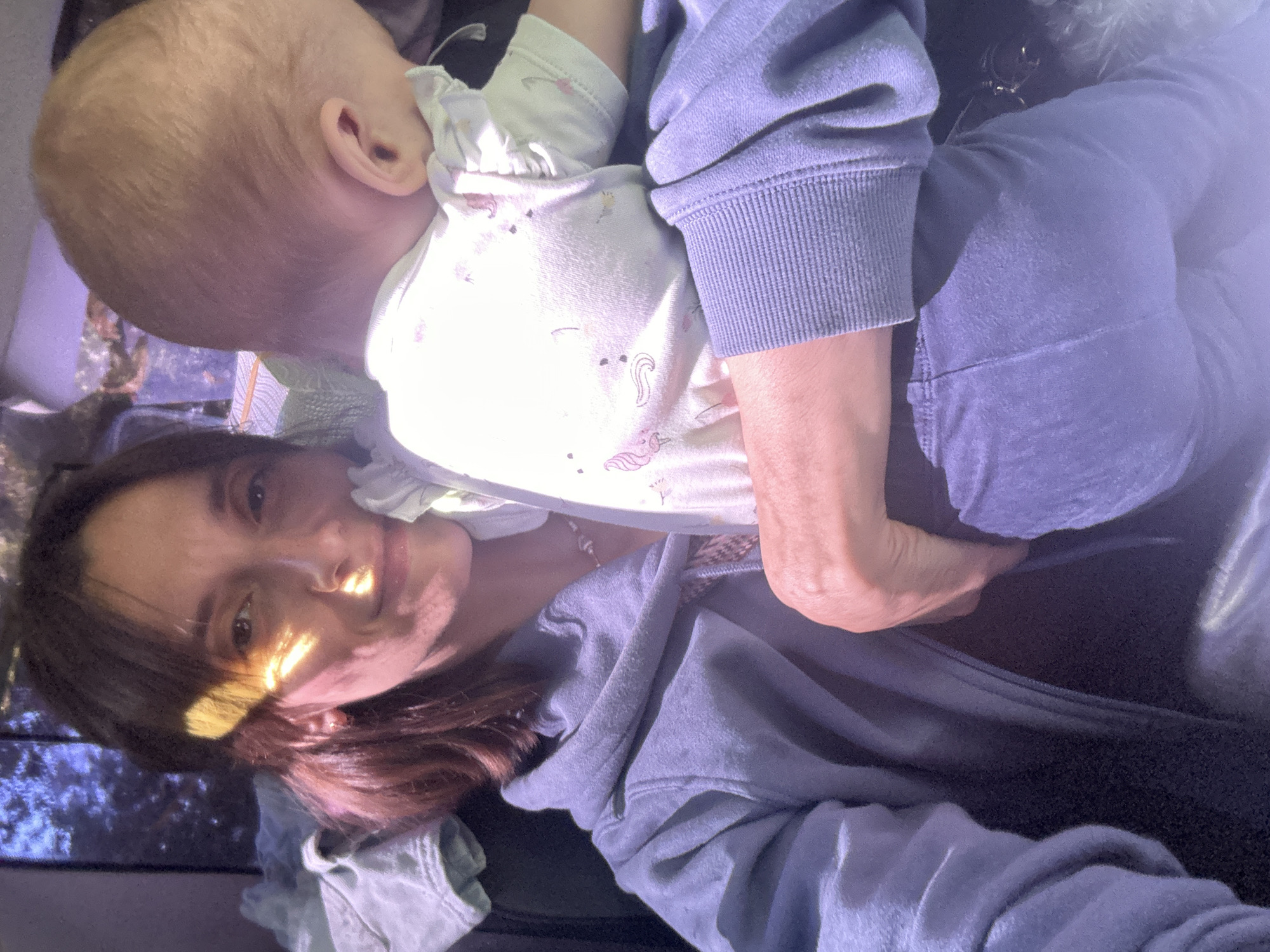 The image displays an adult female holding an infant, both seated in what appears to be an interior space, possibly a vehicle, characterized by a window showing foliage in the background.
The adult female, with medium-length brown hair, is positioned facing forward, slightly angled towards the viewer. A strong diagonal beam of warm, golden light illuminates the left side of her face and hair, creating a bright stripe across her eye and cheek. She exhibits a subtle, contented expression, with light freckling visible on her nose and cheek. She is wearing a blue-grey hooded sweatshirt over a darker undershirt, with a delicate silver-toned necklace visible around her neck. Her left arm supports the infant.
The infant, observed from behind, has light-colored hair and is resting its head against the adult female's shoulder or chest. Its right ear is visible. The infant is dressed in a light-colored, possibly white or cream, short-sleeved top adorned with faint pink and purple patterns, including what appear to be flamingos and unicorns, and features ruffled sleeves. Dark blue or purple pants cover its lower body. The infant appears calm or asleep.
The background shows diffused light through a window, revealing dark green or blue foliage, suggesting an outdoor environment. The interior elements are largely dark and indistinct, with crumpled light-colored fabric visible on a seat to the adult's left. The overall lighting is a mixture of the direct warm sunlight and a cooler, ambient blue-purple tone in the shaded areas and background.