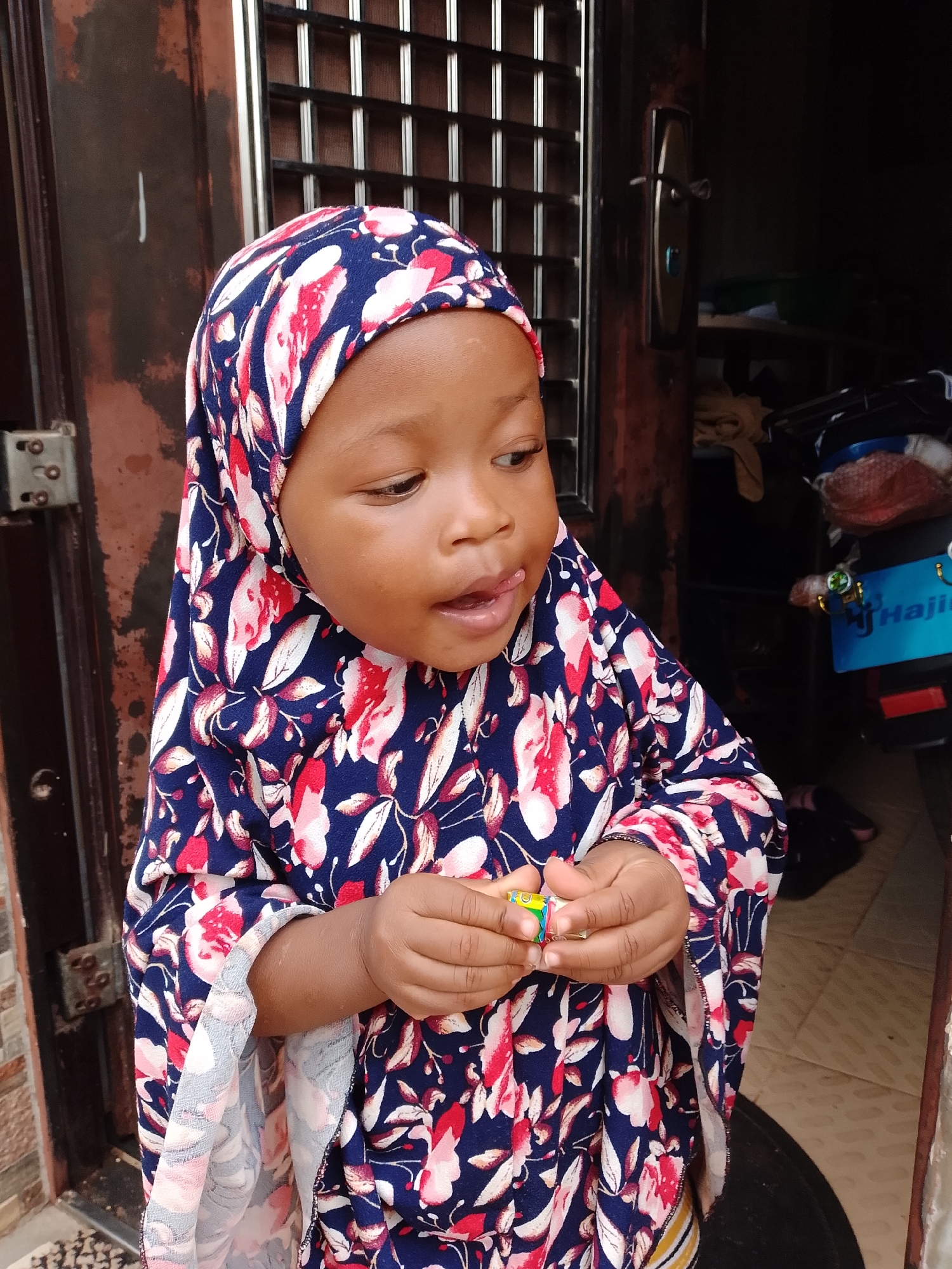 A young child, wearing a blue and red floral hijab and dress, stands in front of a dark brown door. The child is looking to the right with their tongue slightly out, holding a small, colorful object in their hands. To the left of the child, a metal gate is visible, and to the right, a motorcycle is partially visible with a blue license plate that has white lettering. The background appears to be an outdoor setting, possibly a doorway or entrance in Jos, Nigeria.