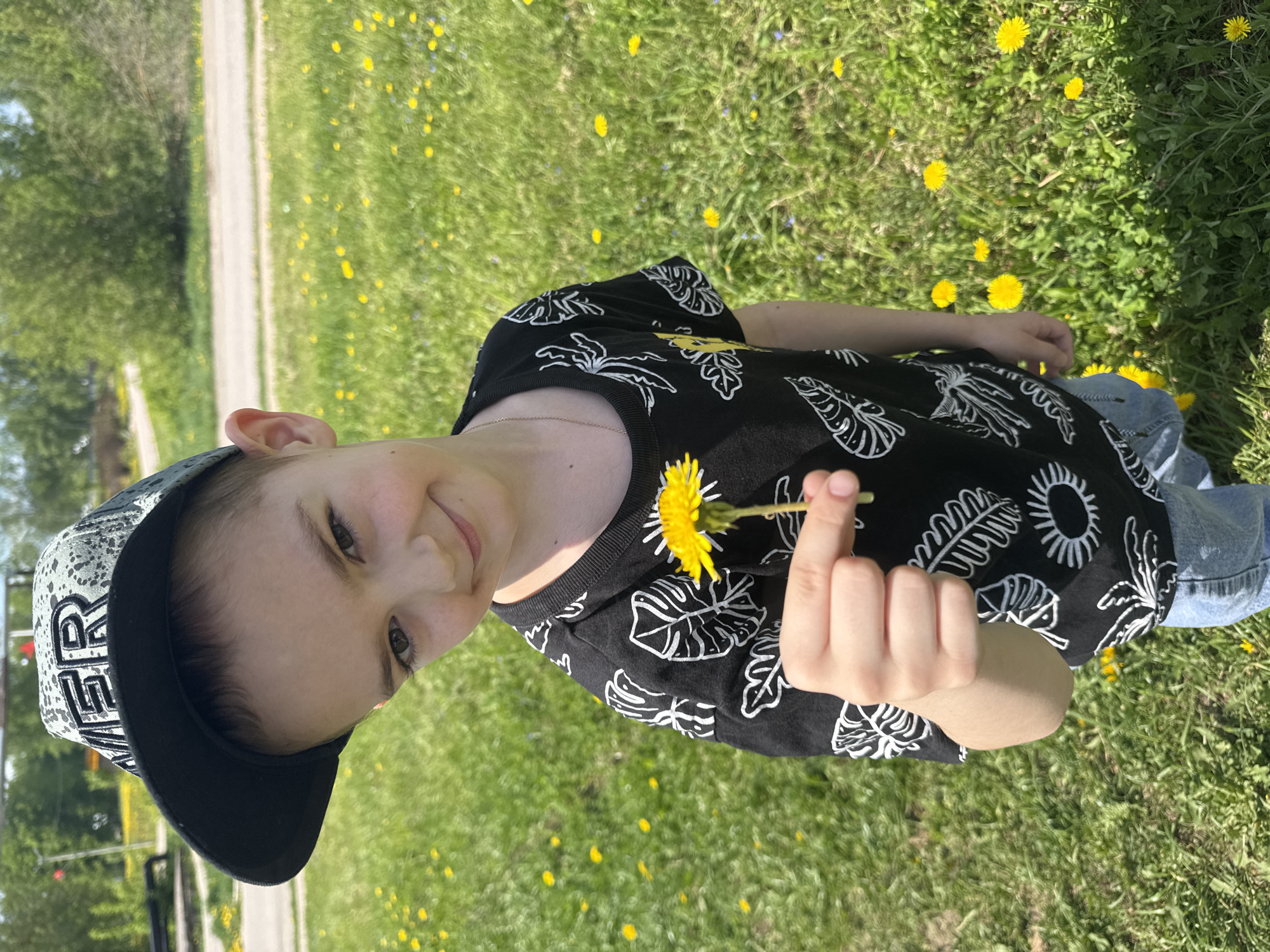 A young child stands in a grassy field filled with dandelions. The child is wearing a black t-shirt with white leaf and sun designs, blue jeans, and a black cap with a white design on the front. The child is holding a yellow dandelion in their right hand and smiling slightly. The background shows a grassy area and a path or road bordered by trees. The photo was taken in Shevlyakovo, Russia.